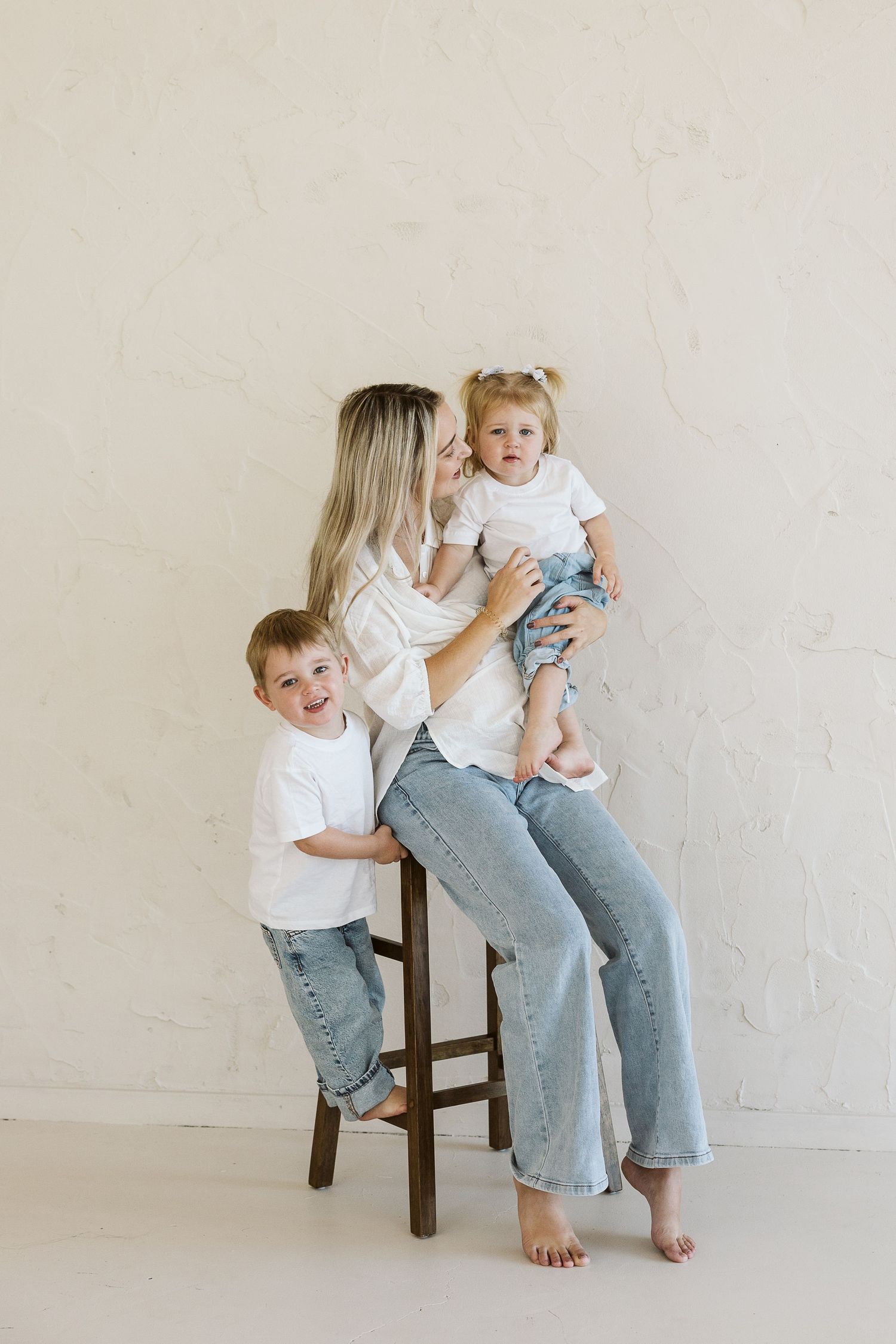 A mother sits on a wooden stool holding her toddler daughter while her young son leans against her, all wearing white tees and jeans.