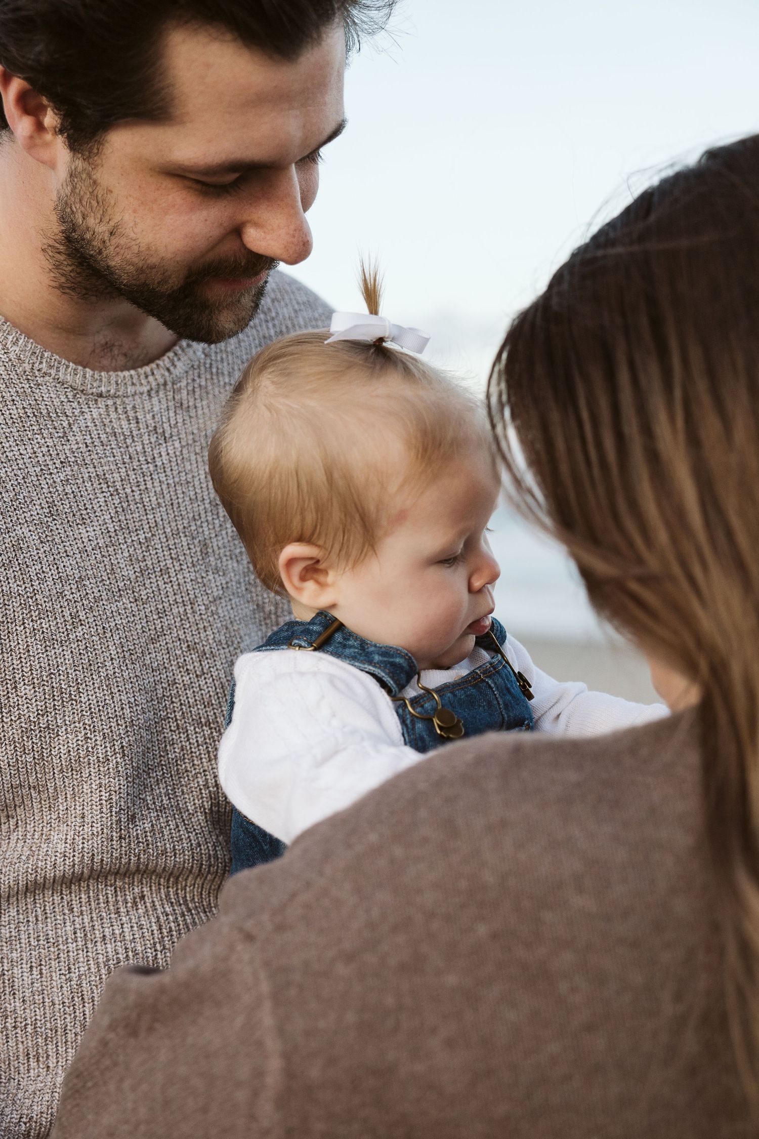 A young family with a bearded father and mother lovingly gazing at their baby girl in denim overalls outdoors.