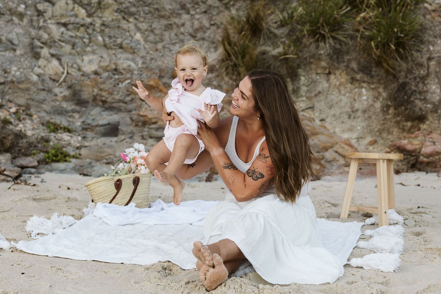 A mother sits on a white blanket at the beach, joyfully lifting her laughing baby in a pink outfit.