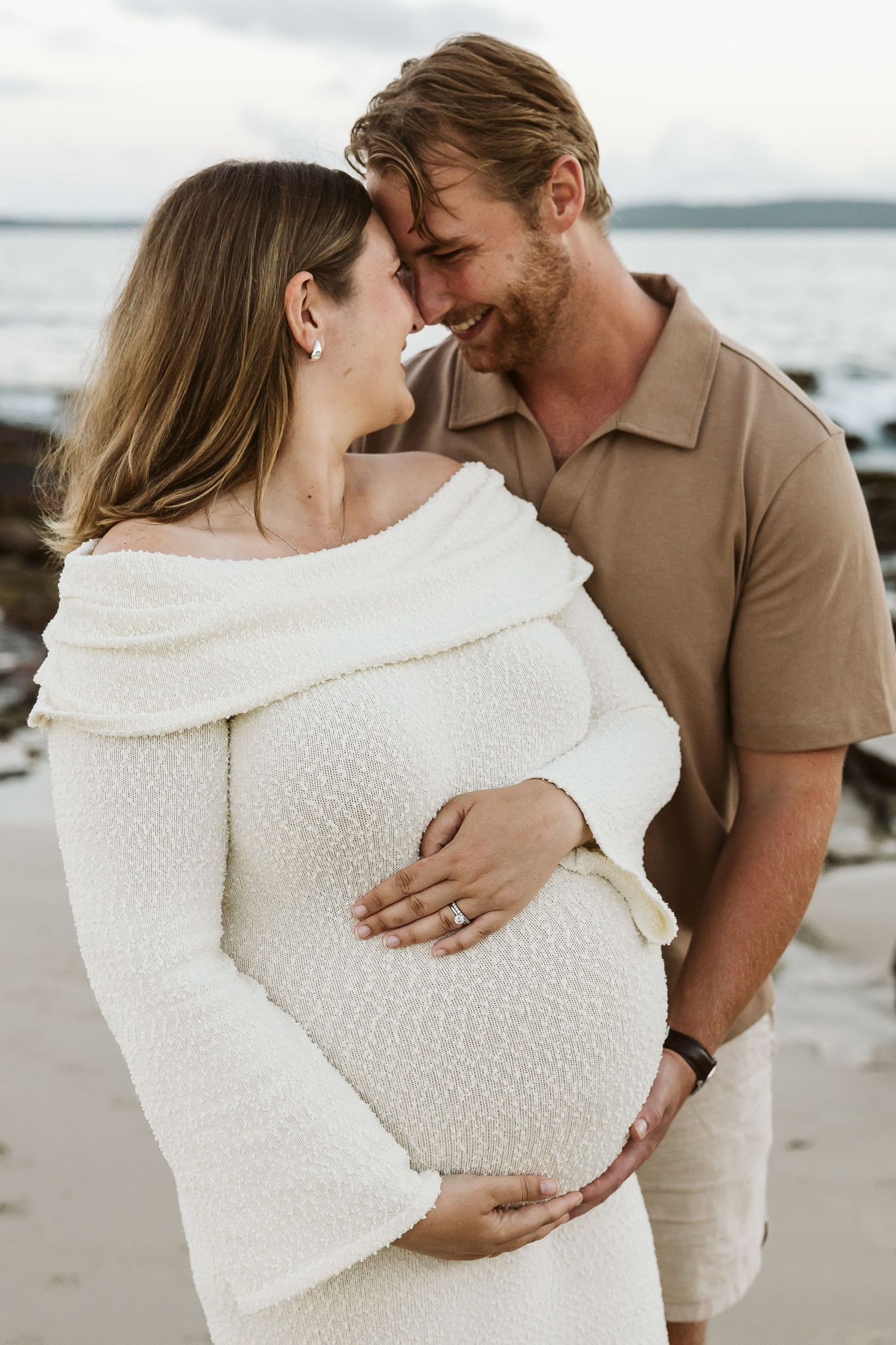 Couple shares forehead touch on beach, man in tan polo embraces pregnant woman in white textured dress.