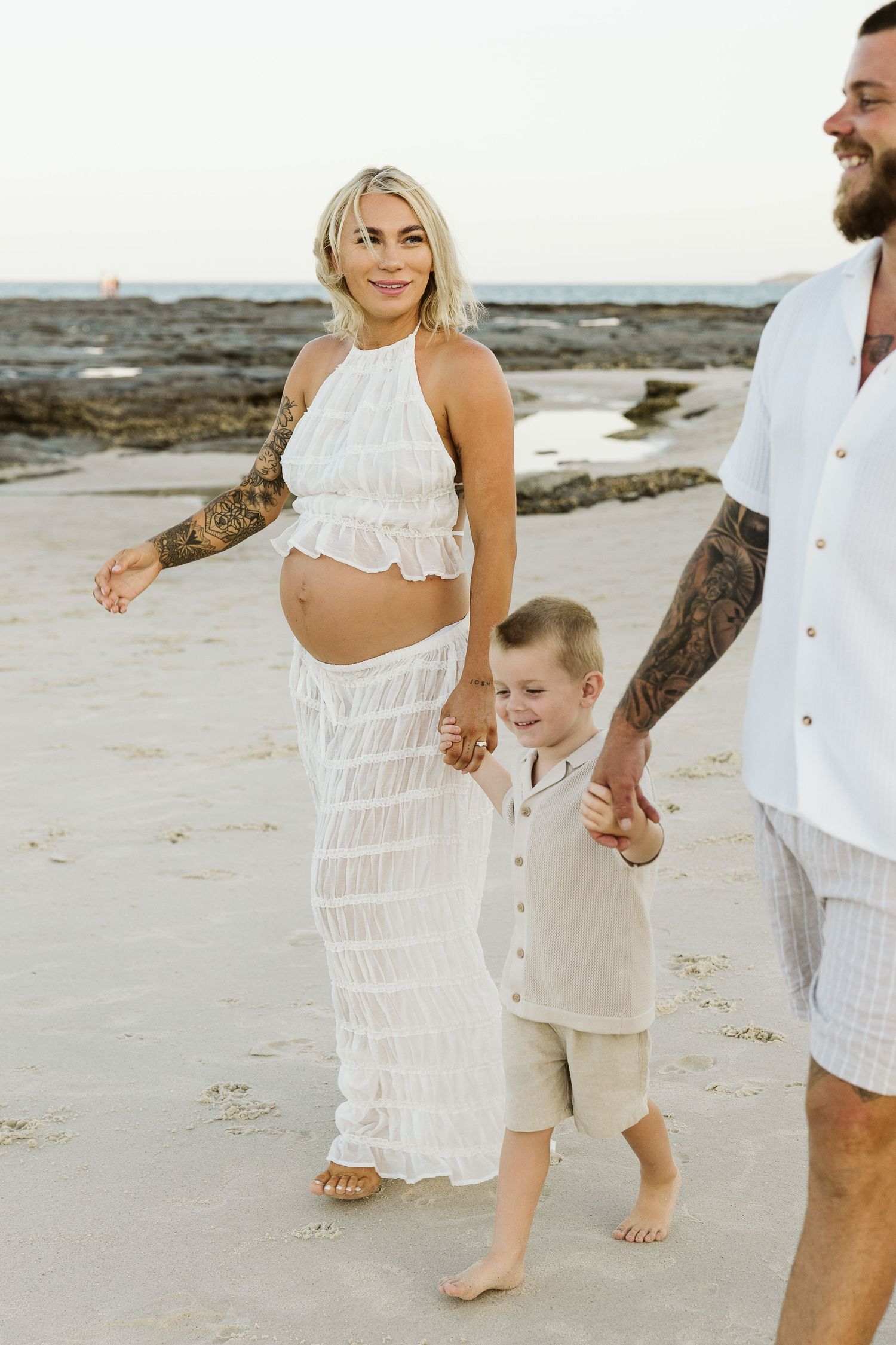 Smiling pregnant blonde woman in white crop top and skirt walking on beach with family.