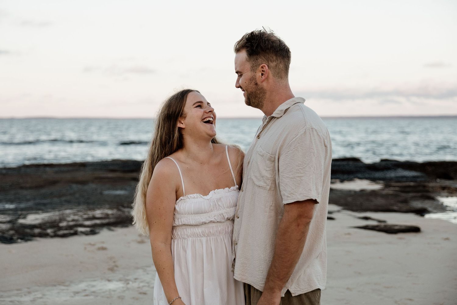 Laughing couple embracing on a sandy beach at sunset, woman in white sundress and man in linen shirt facing each other.