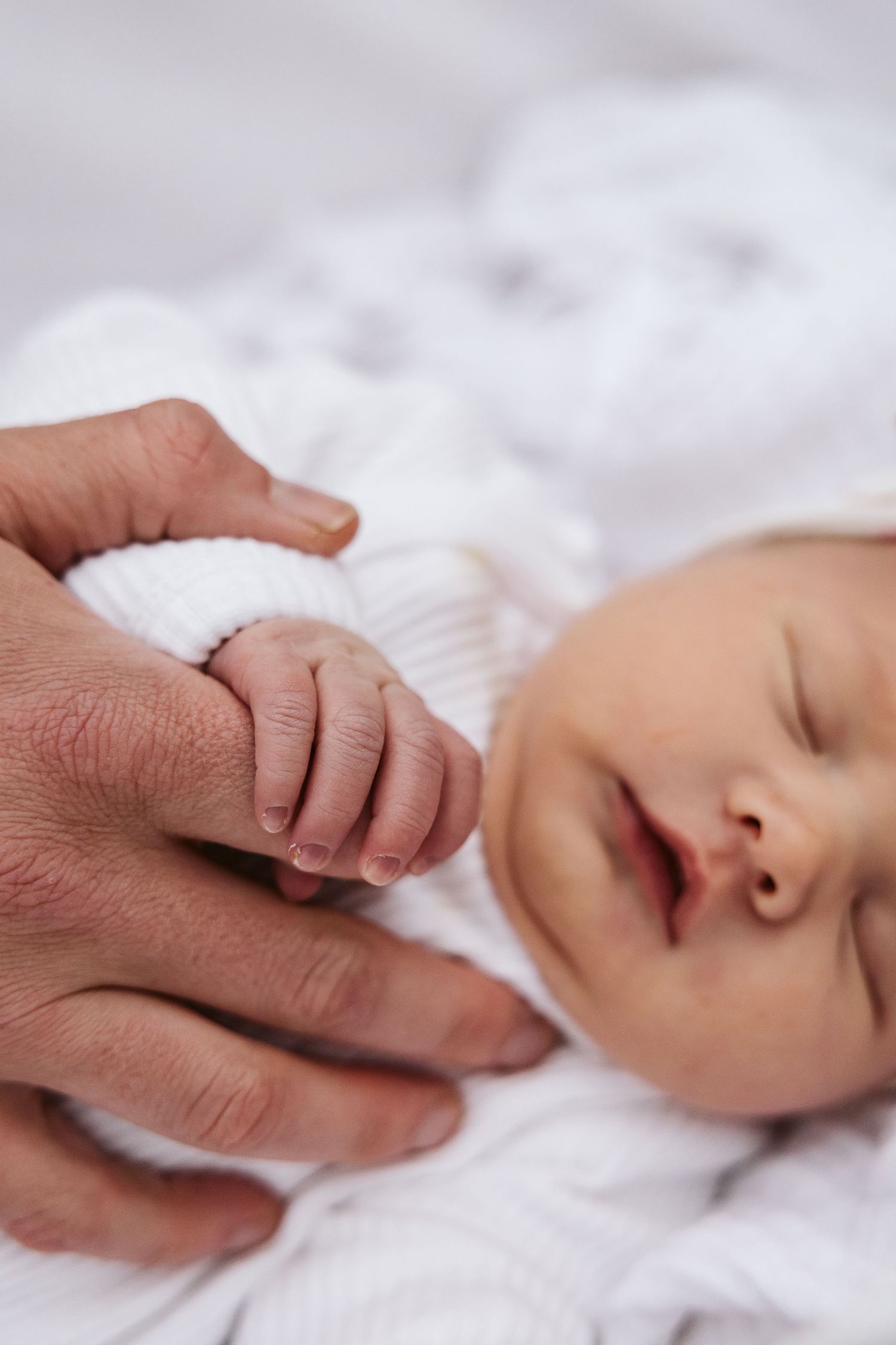 Sleeping newborn baby in white outfit gently gripping an adult finger.