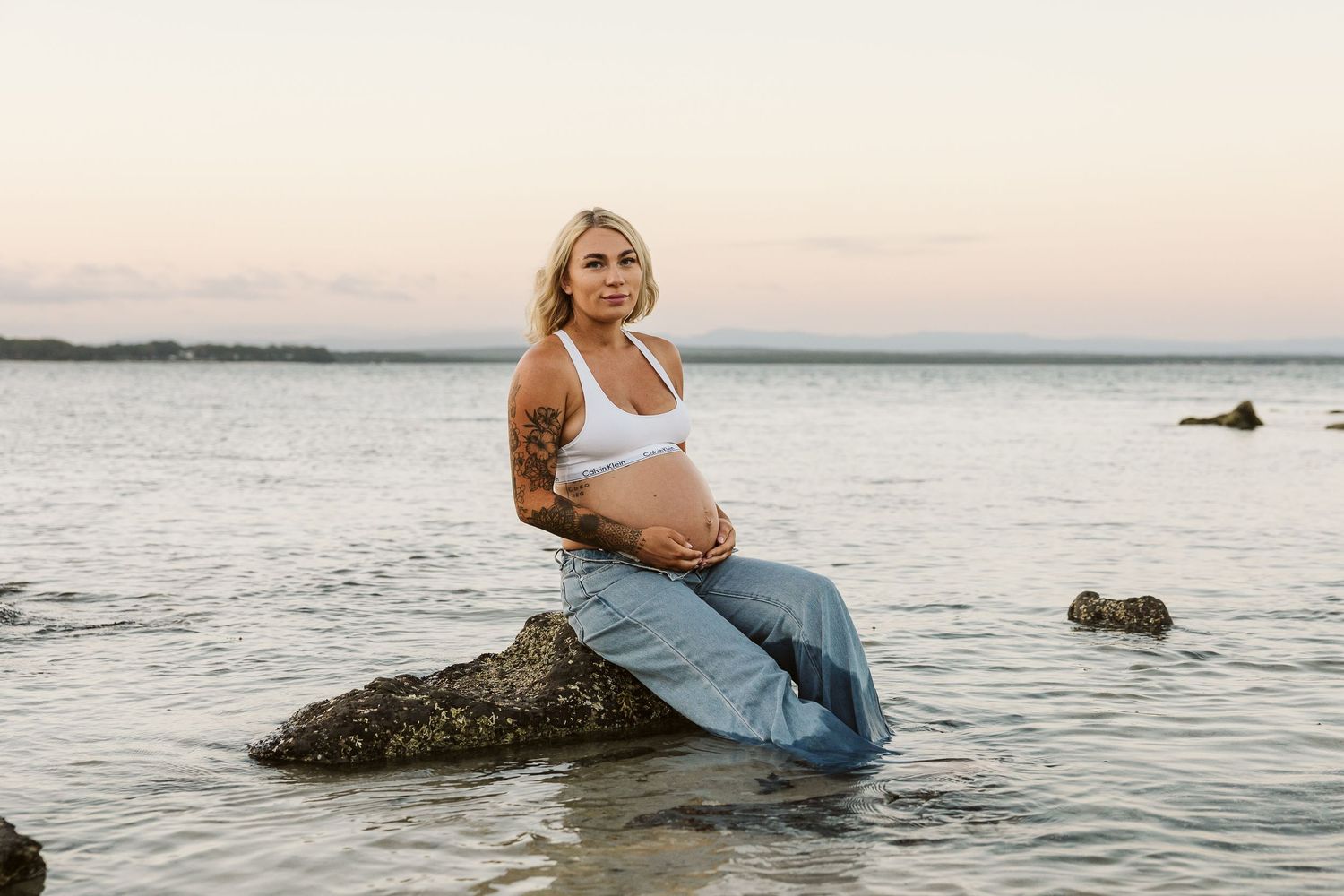 Tattooed pregnant woman sitting on coastal rocks at sunset cradling her baby bump.