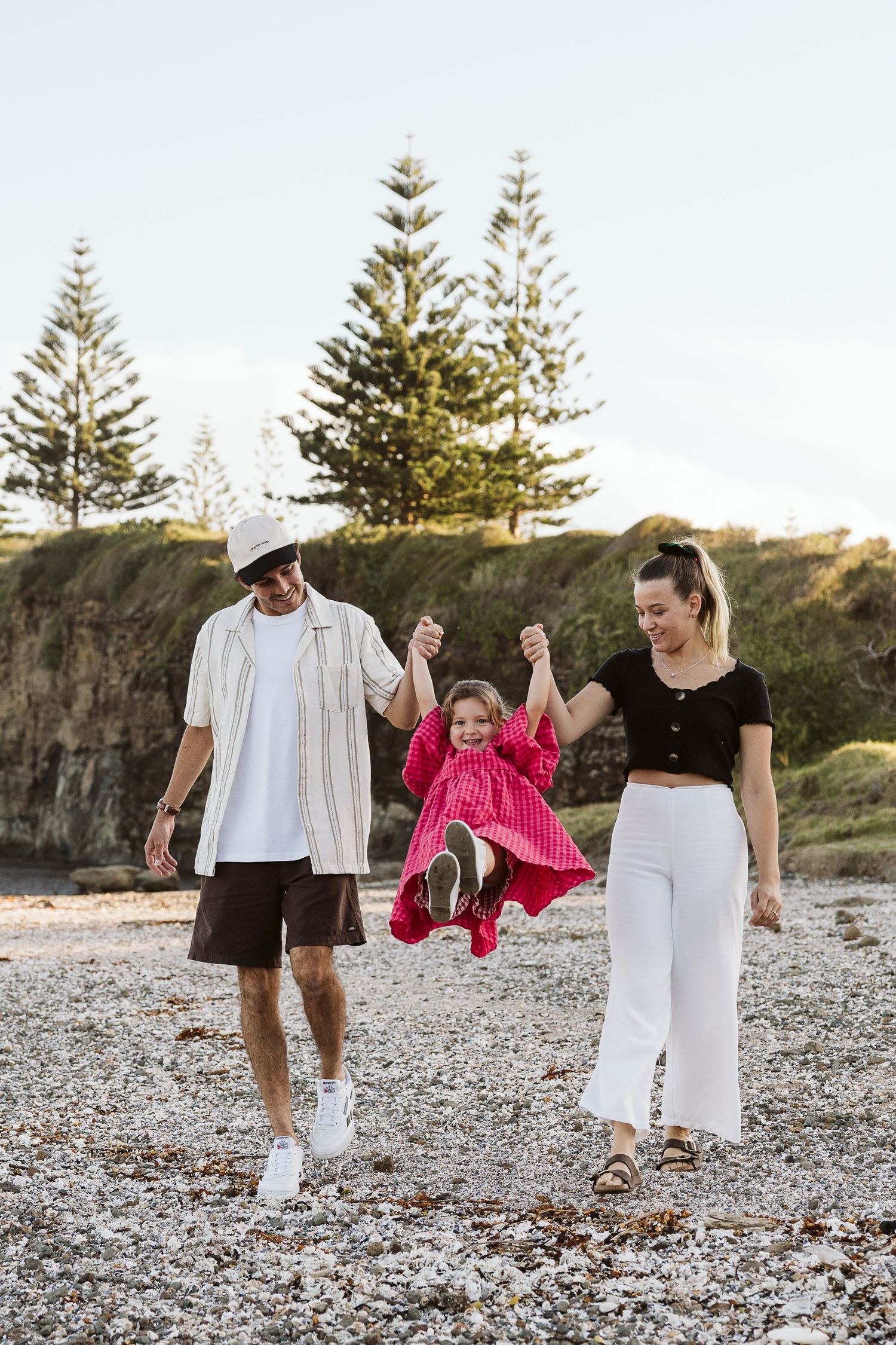 Family of three enjoying a sunny day on a rocky beach, parents swinging their young daughter in a pink dress.