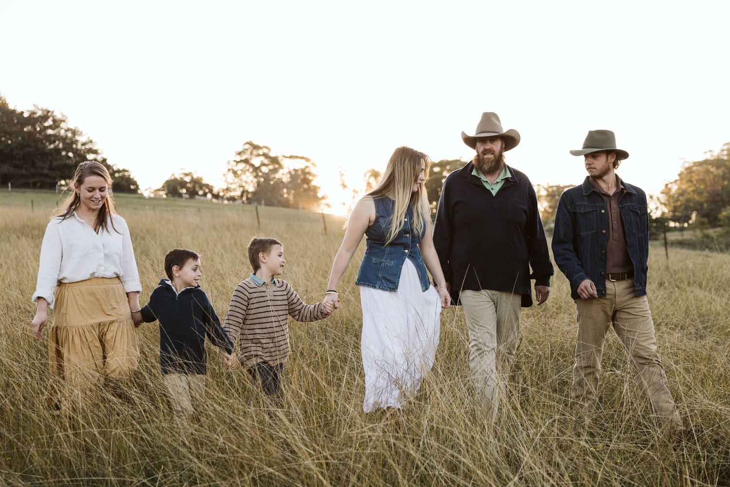 Family of six walking through golden grass field at sunset, wearing casual country attire including cowboy hats.