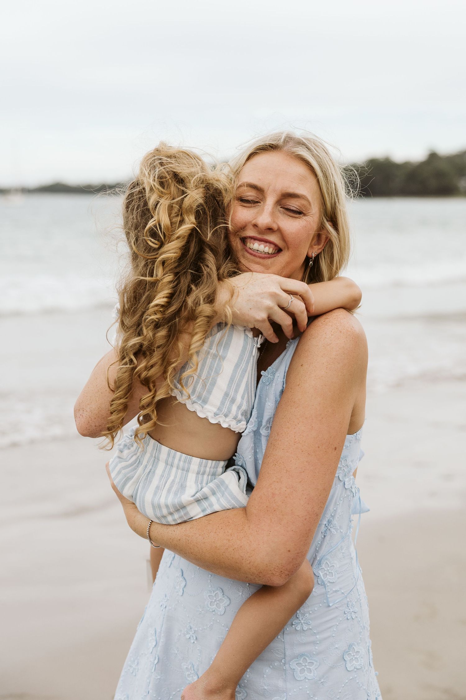 Smiling blonde mother hugging curly-haired daughter on beach, wearing light blue dresses on overcast day.