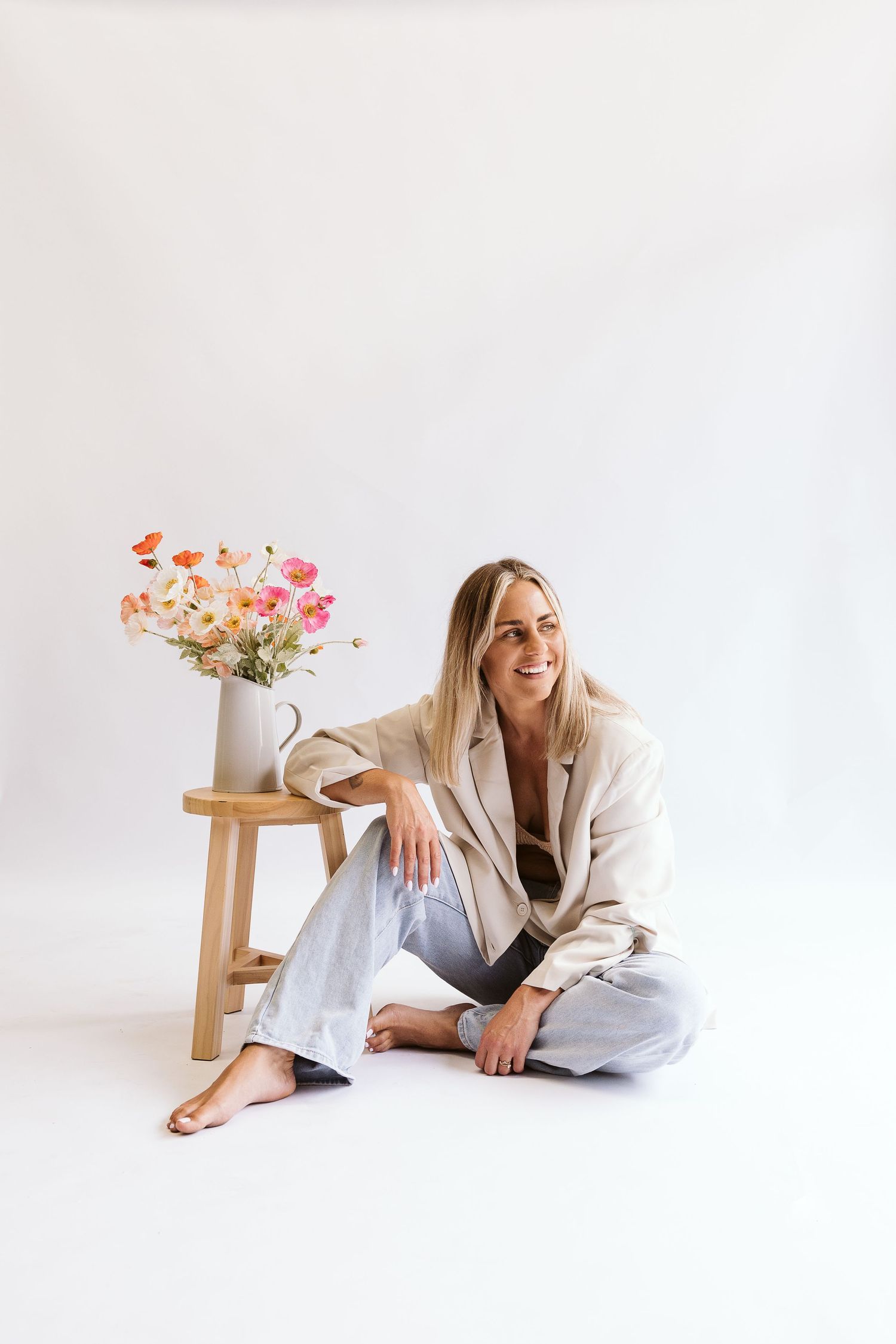 Blonde woman sitting barefoot on white studio floor beside wooden stool with colorful flower bouquet.