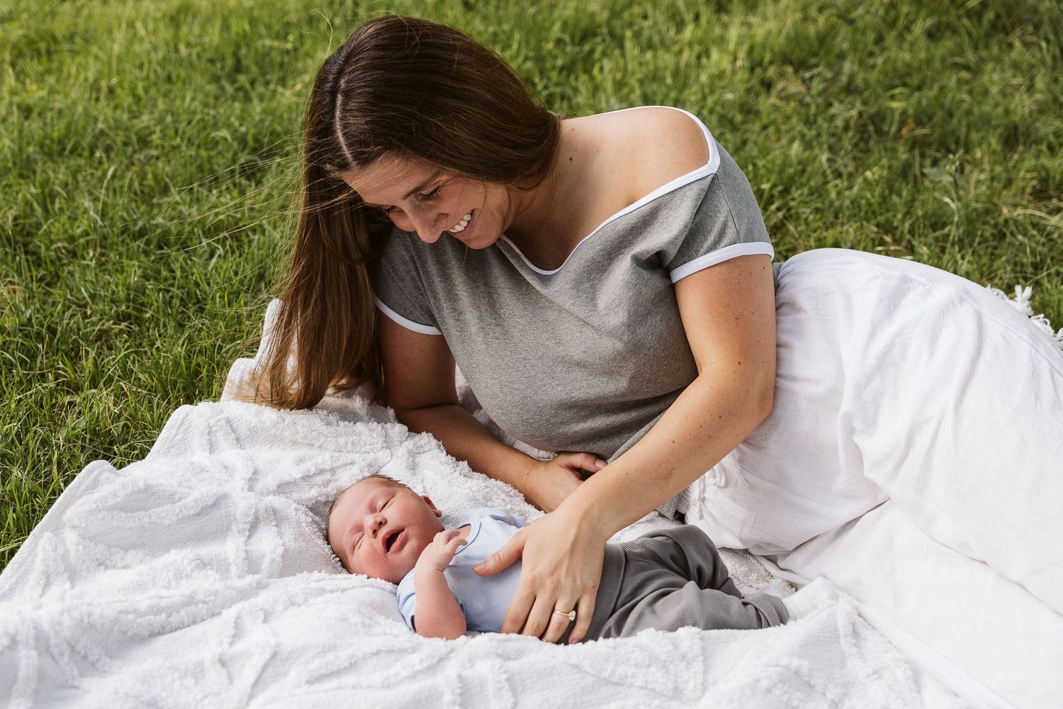 Smiling mother sitting on grass with newborn baby on white blanket outdoors.