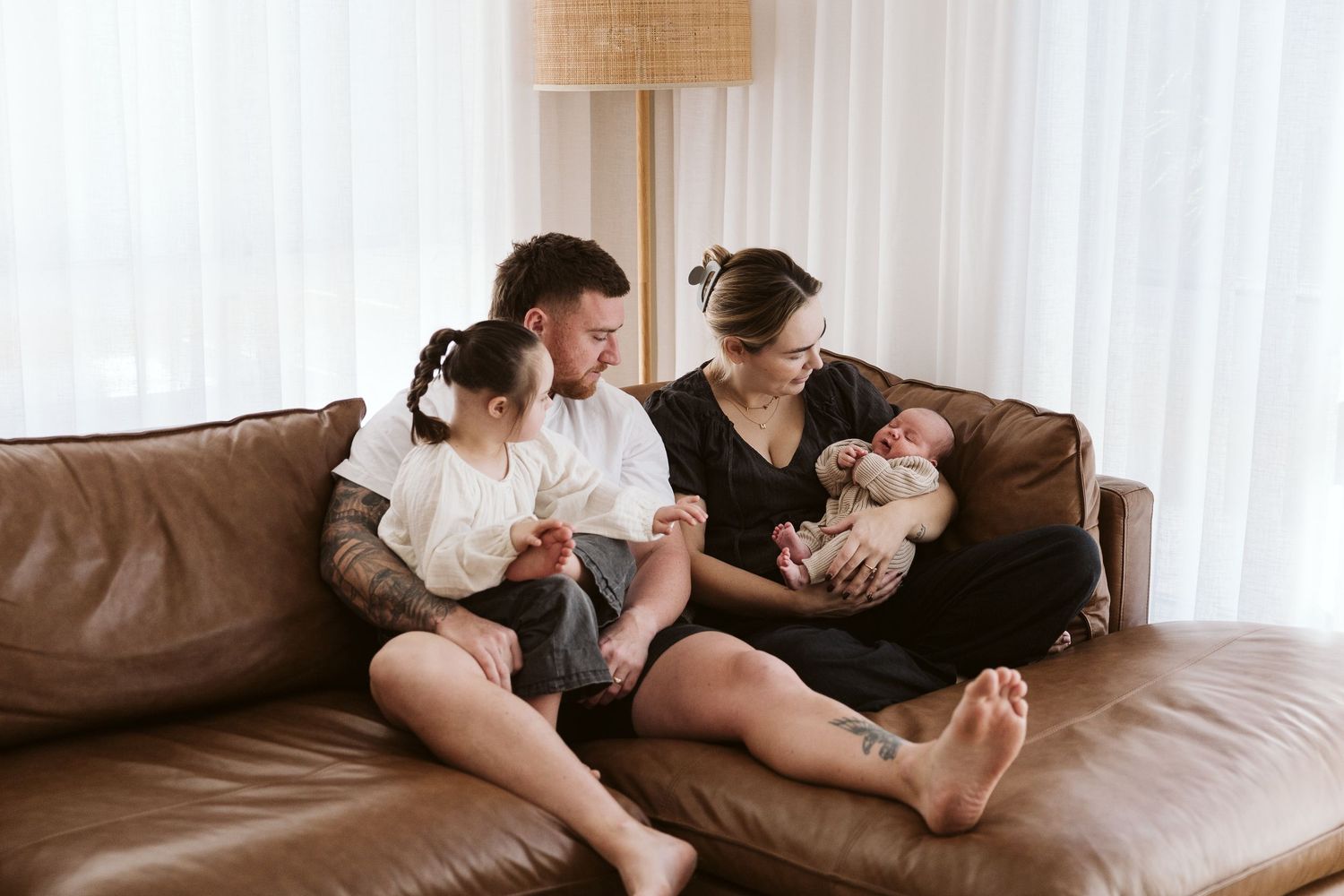 Family of four relaxing on brown leather sofa, mother holding newborn while toddler sits with father.