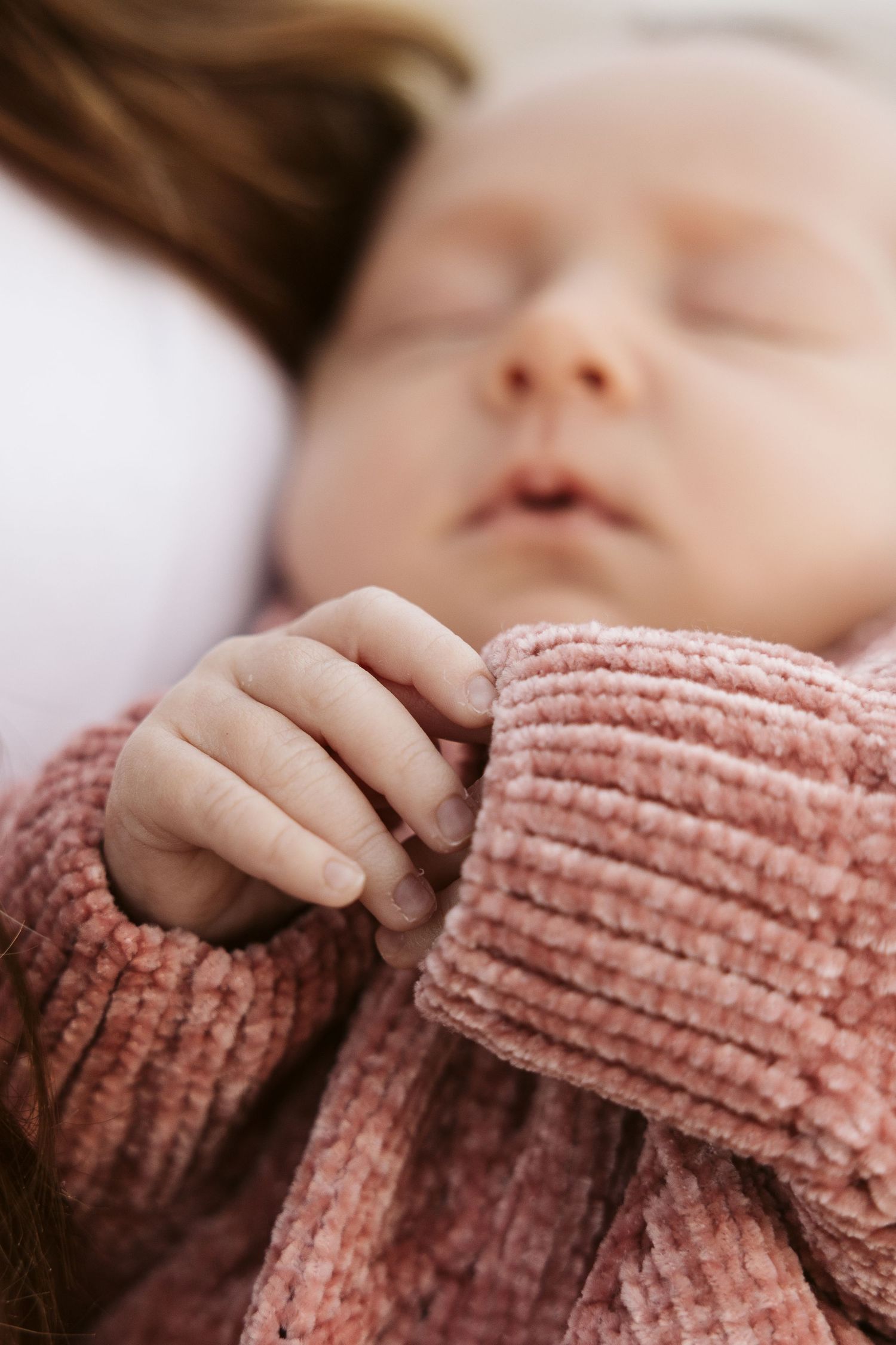 Close-up of sleeping newborn baby in pink chenille sweater with tiny hand visible.