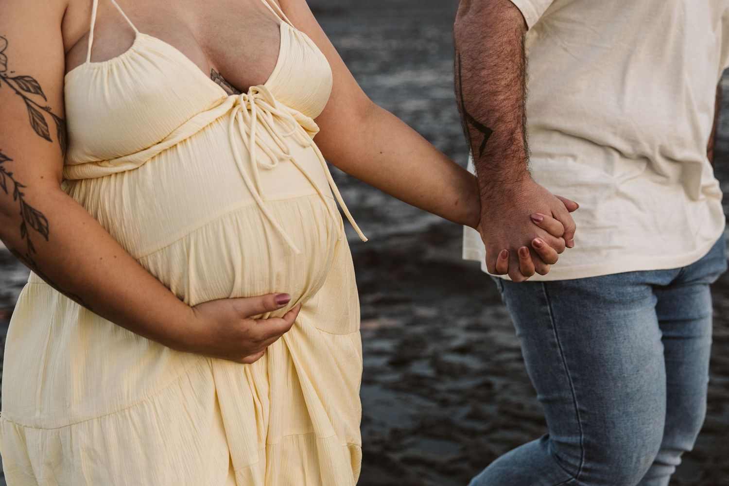 Close-up of pregnant woman in yellow dress holding belly while partner holds her hand.