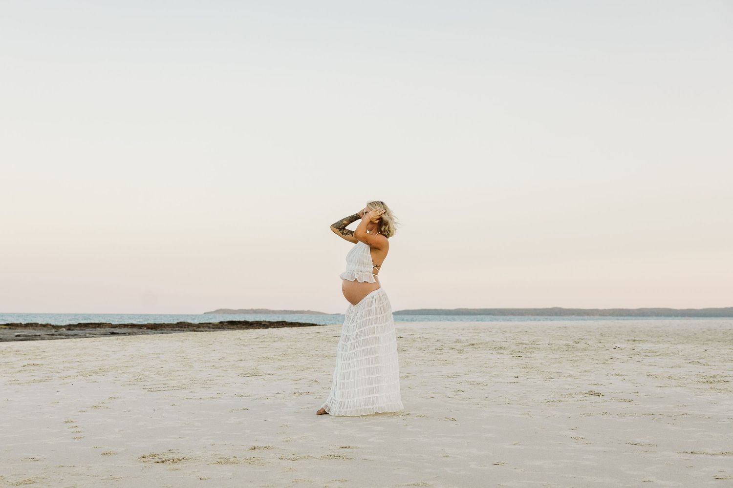 Pregnant blonde woman in white crop top and striped maxi skirt posing on open sandy beach at golden hour.