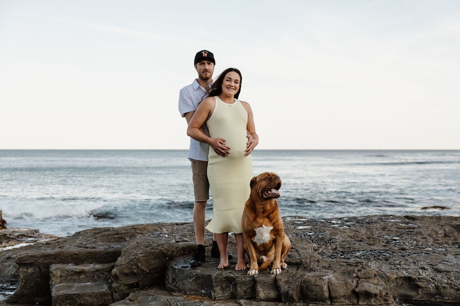 Pregnant couple and brown Mastiff dog pose on rocky coastal shore at sunset.
