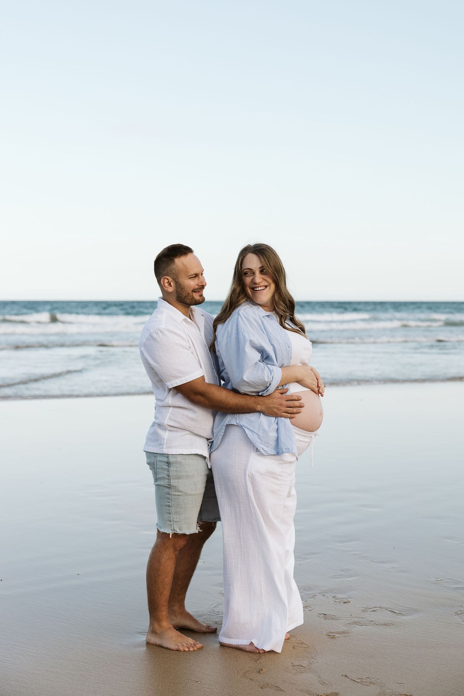 Happy couple embracing on beach at sunset, pregnant woman in blue shirt and white skirt smiling.