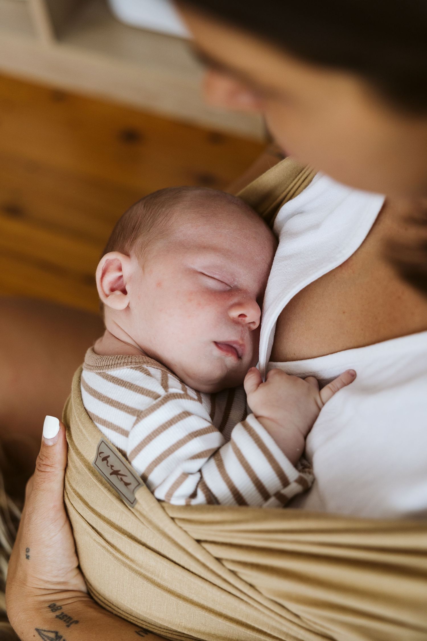 Sleeping newborn baby in striped onesie cradled against mother in a tan wrap carrier.