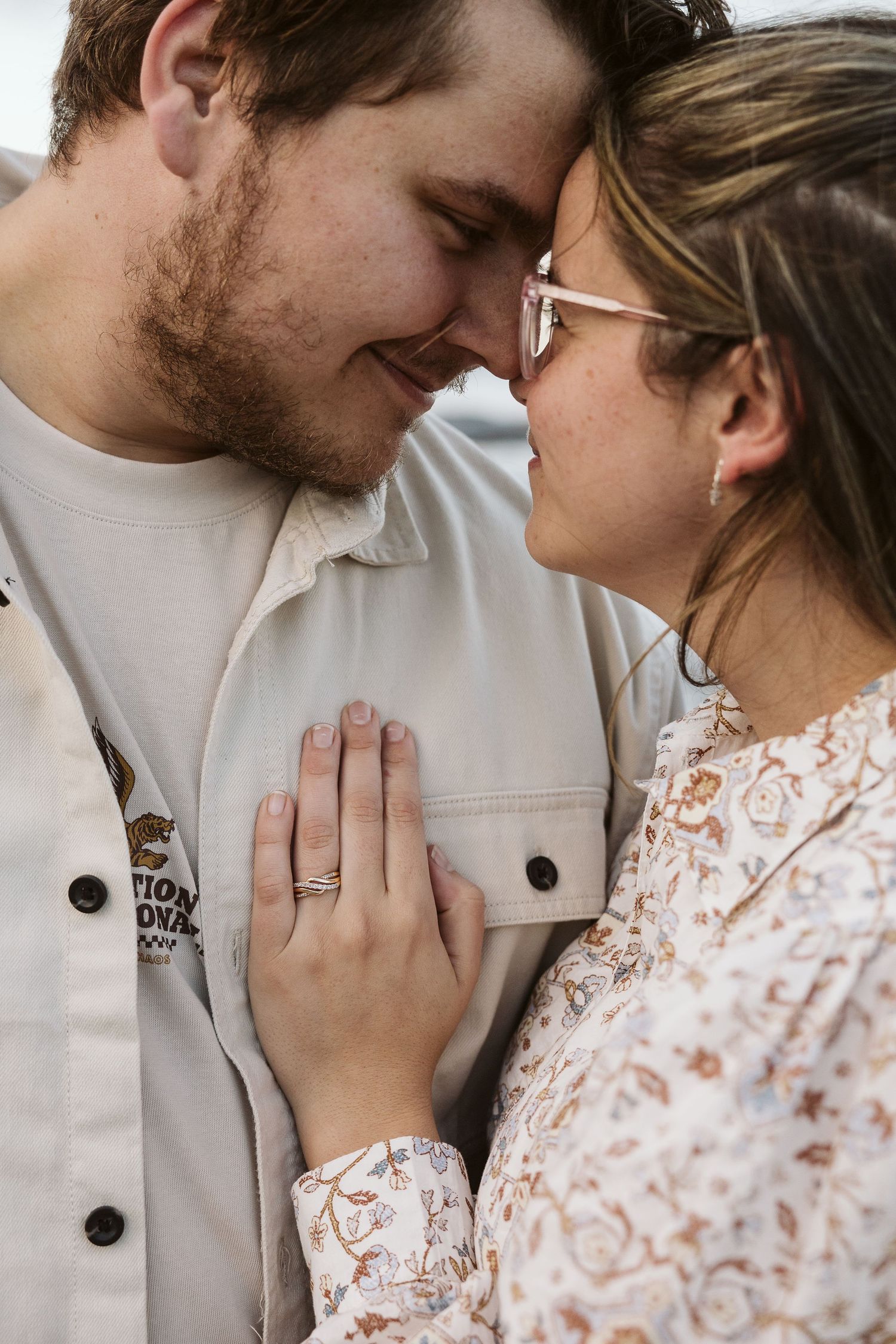 Engaged couple touching foreheads and smiling, woman showing diamond ring on mans chest outdoors.