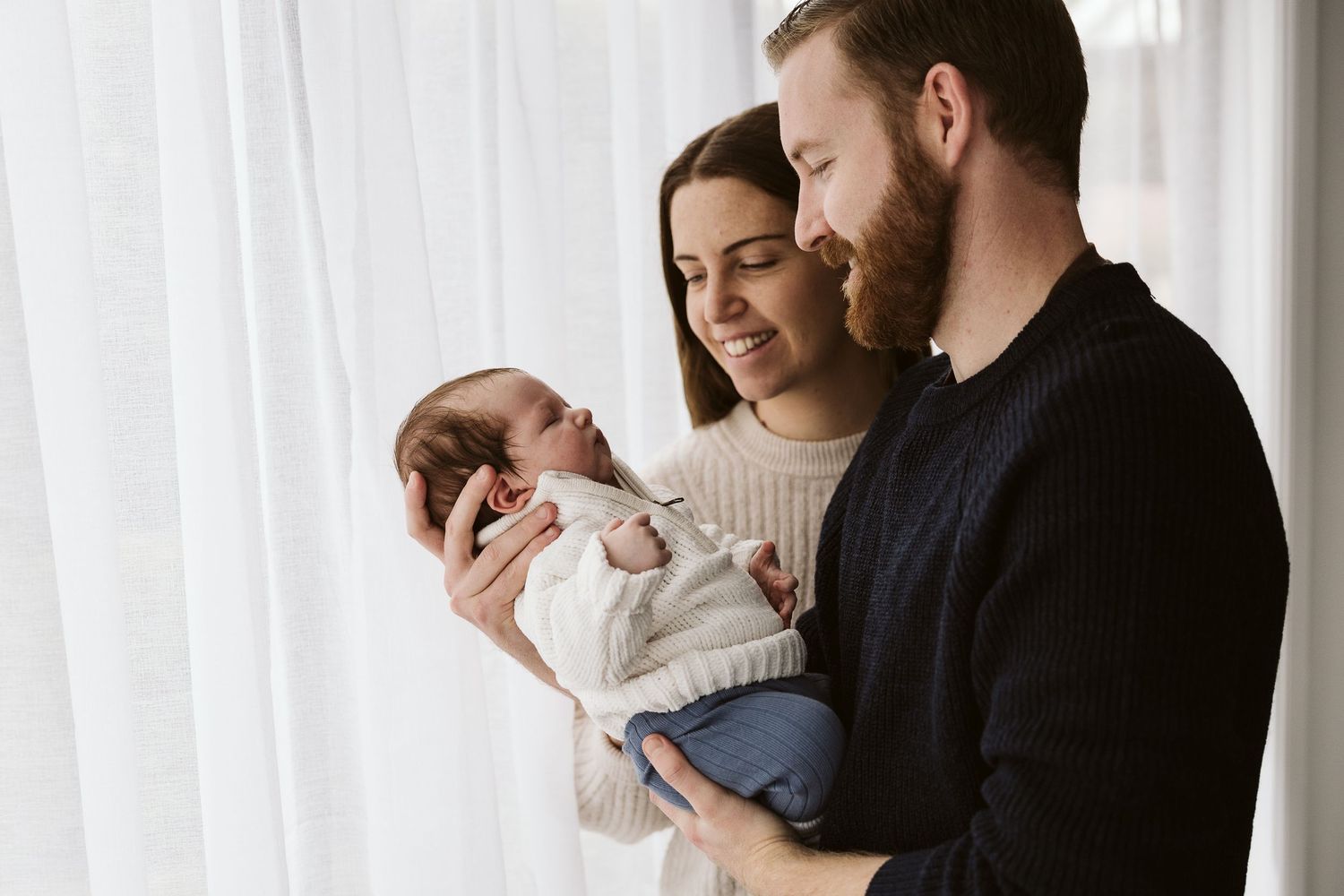 Happy couple smiling at their newborn baby by a bright window.