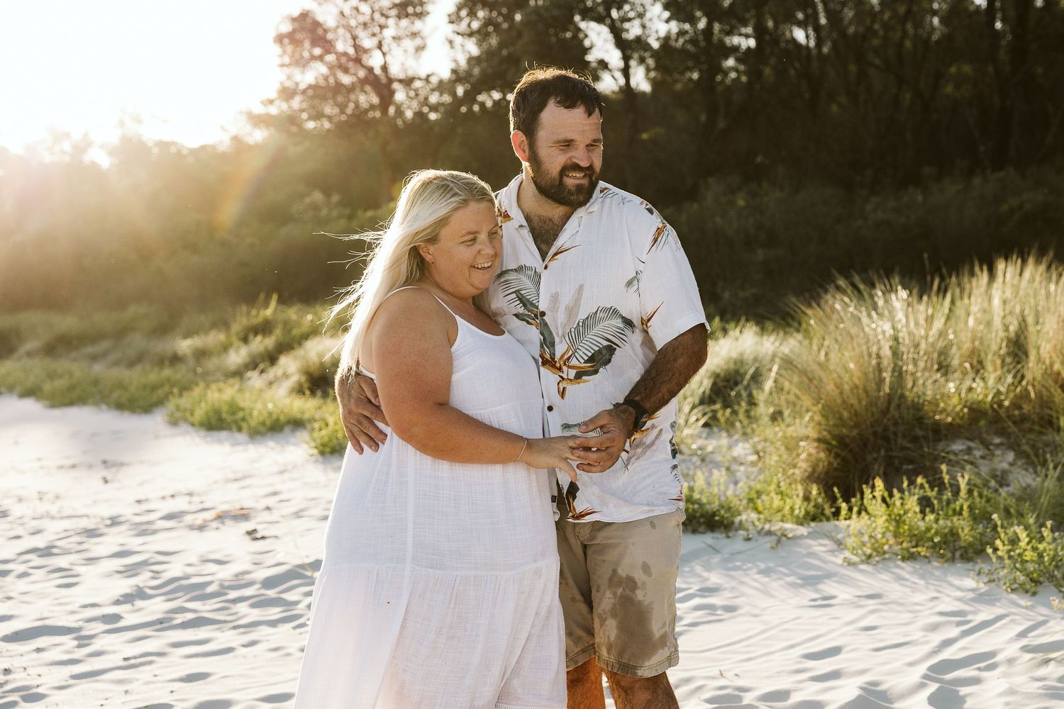 Smiling couple holding hands on sandy beach in warm sunset light.