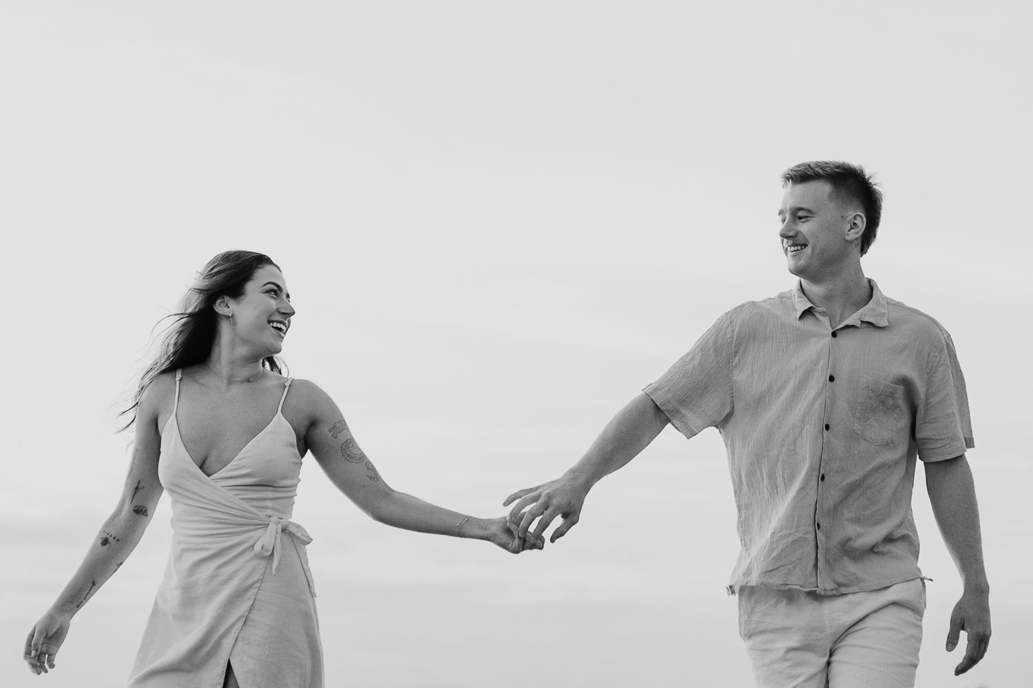 Black and white photo of a smiling couple holding hands and walking together on a breezy beach.