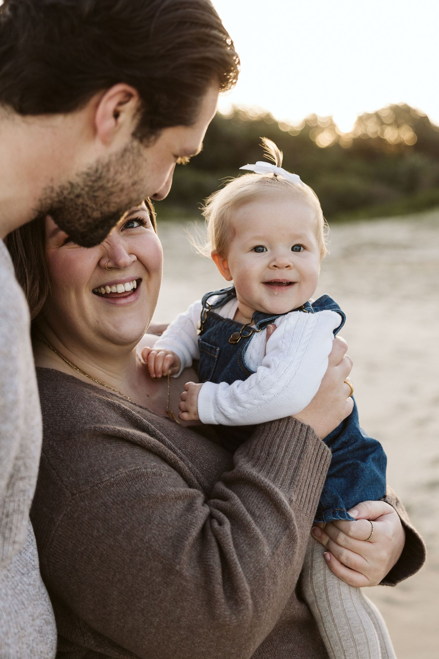 A smiling couple holds their happy baby girl on a sunny beach, capturing a joyful family moment outdoors.
