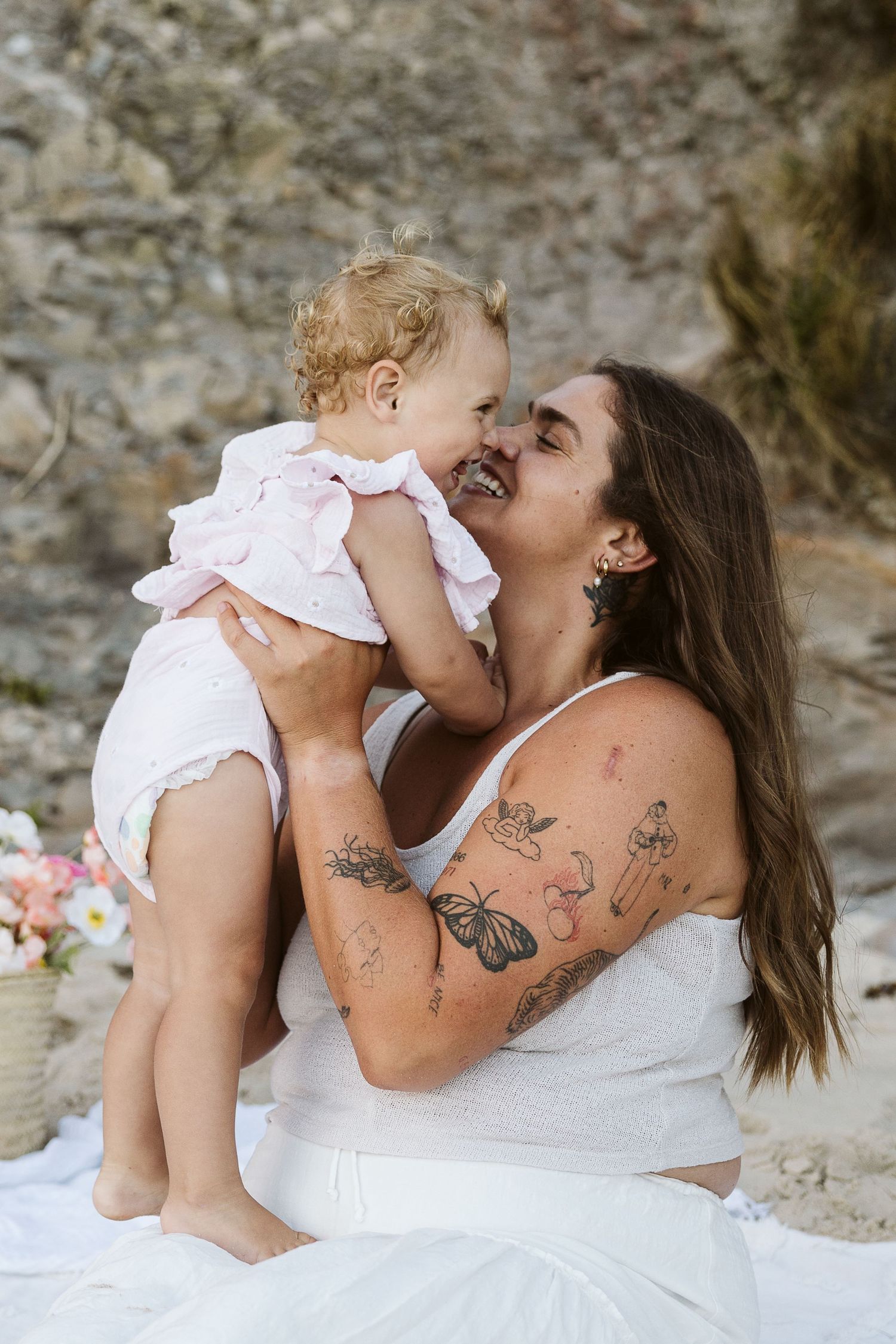Tattooed mother joyfully holds laughing baby girl in pink dress during outdoor photo session.