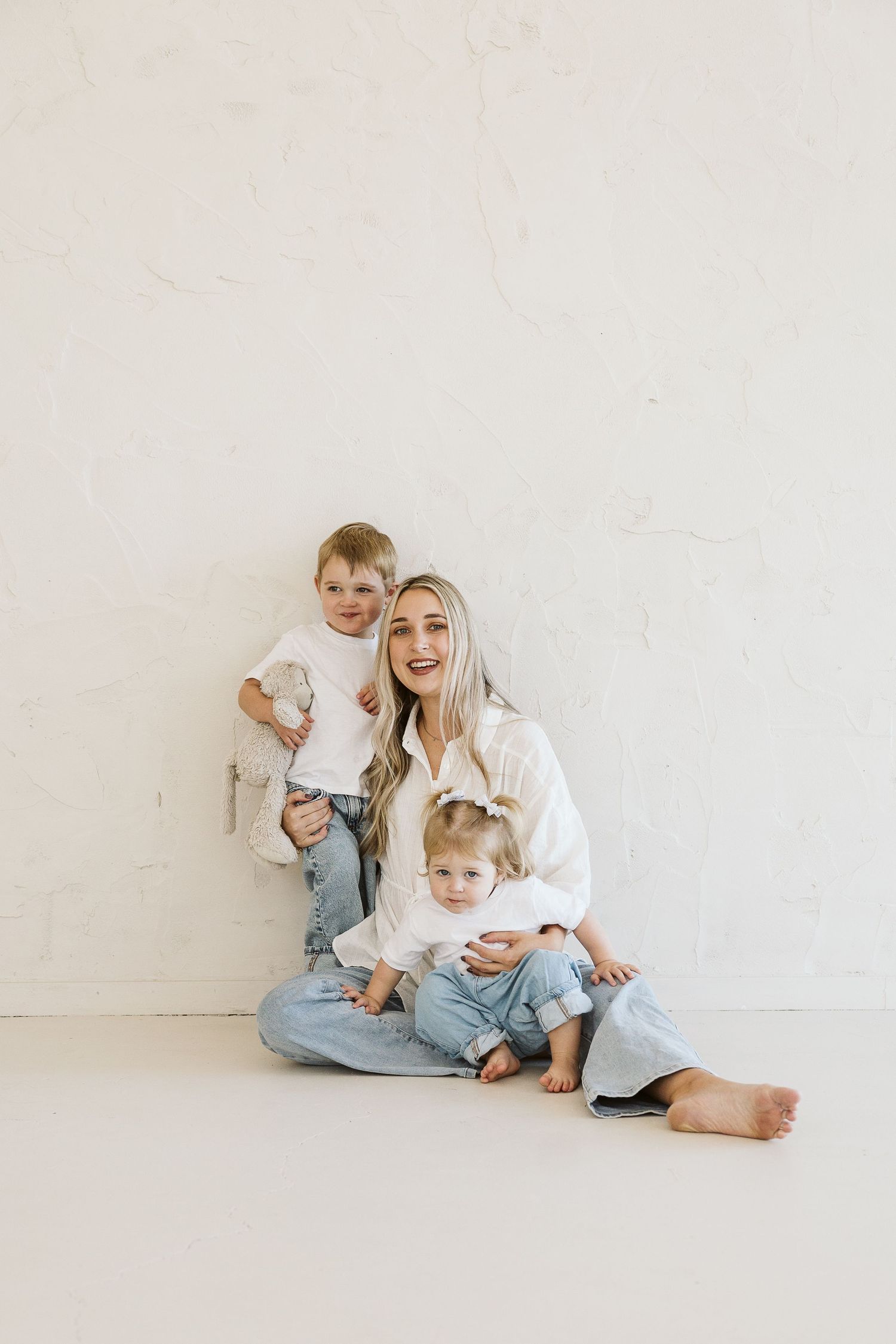 A smiling woman in white shirt and jeans sits on floor with two young children in a bright white studio.
