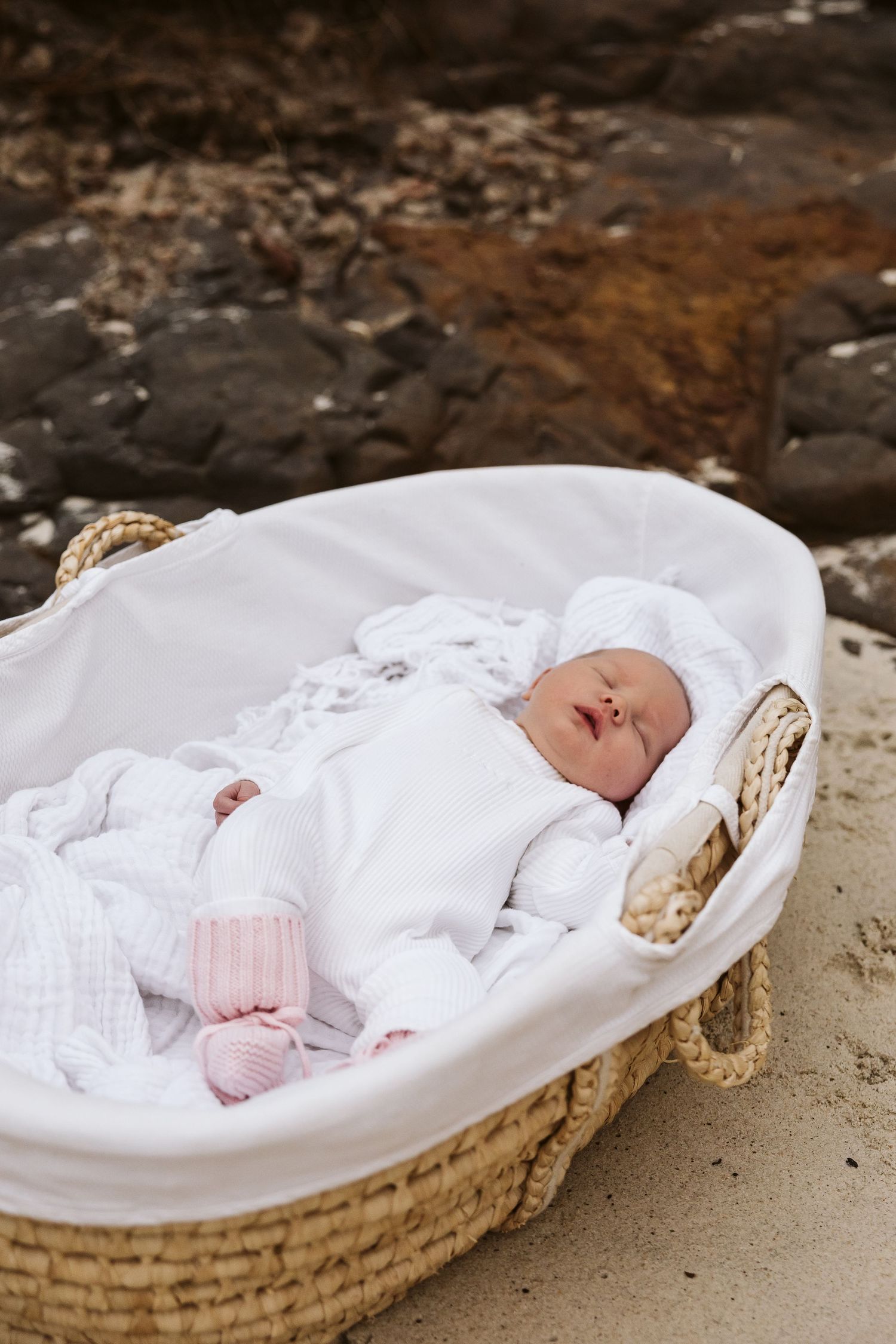 Sleeping newborn baby dressed in white lies in wicker Moses basket on beach.