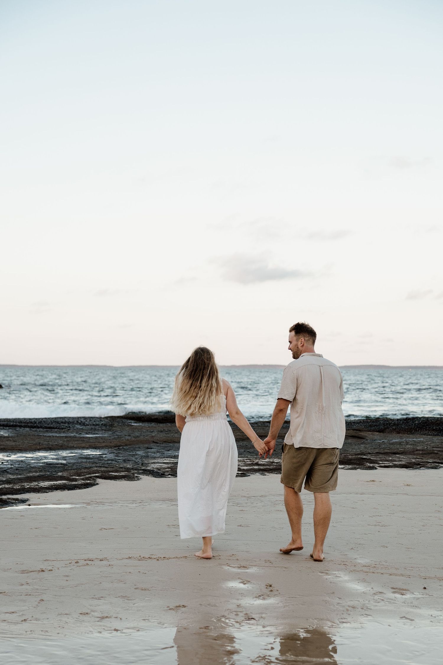 Couple walking hand in hand toward the ocean at sunset, woman in flowing white dress and man in beige shirt and shorts.
