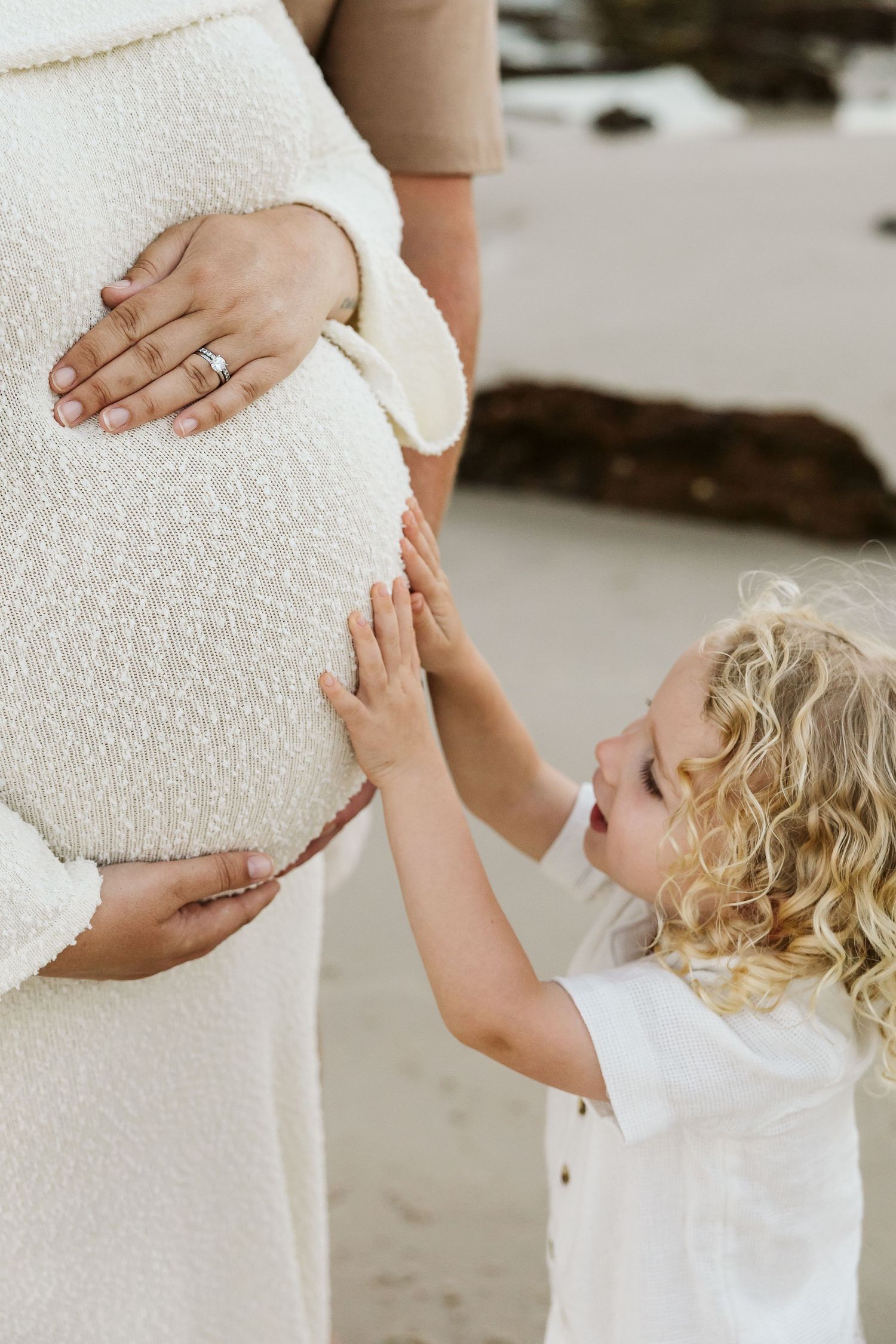 Curly-haired toddler touches pregnant mothers baby bump in cream knit dress on sandy beach.