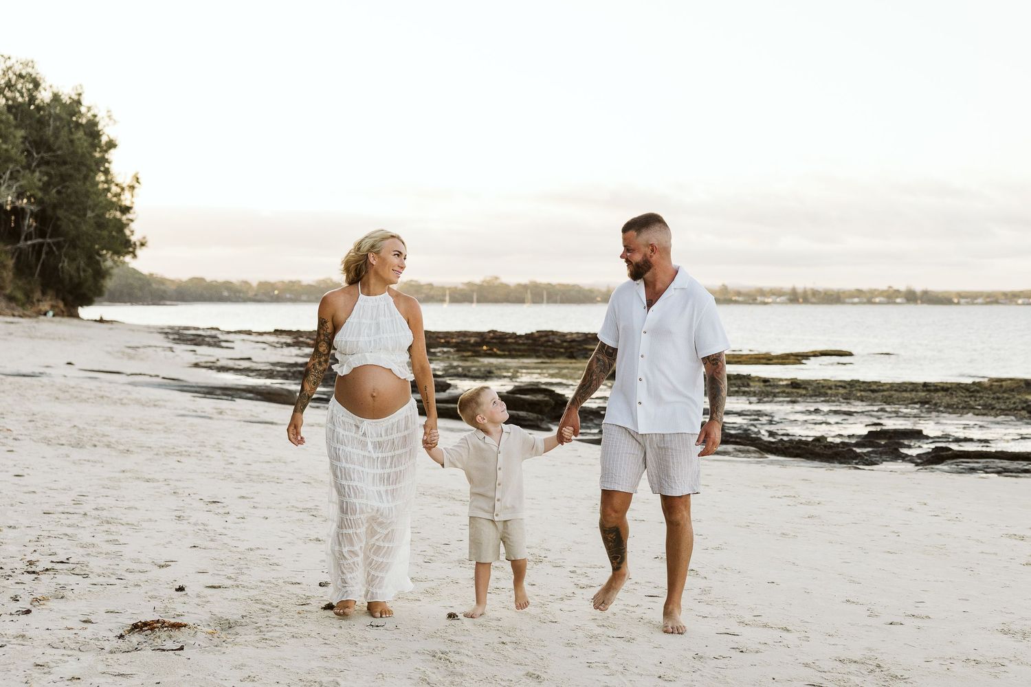 Family maternity photo on beach with pregnant woman, man, and toddler in white outfits.