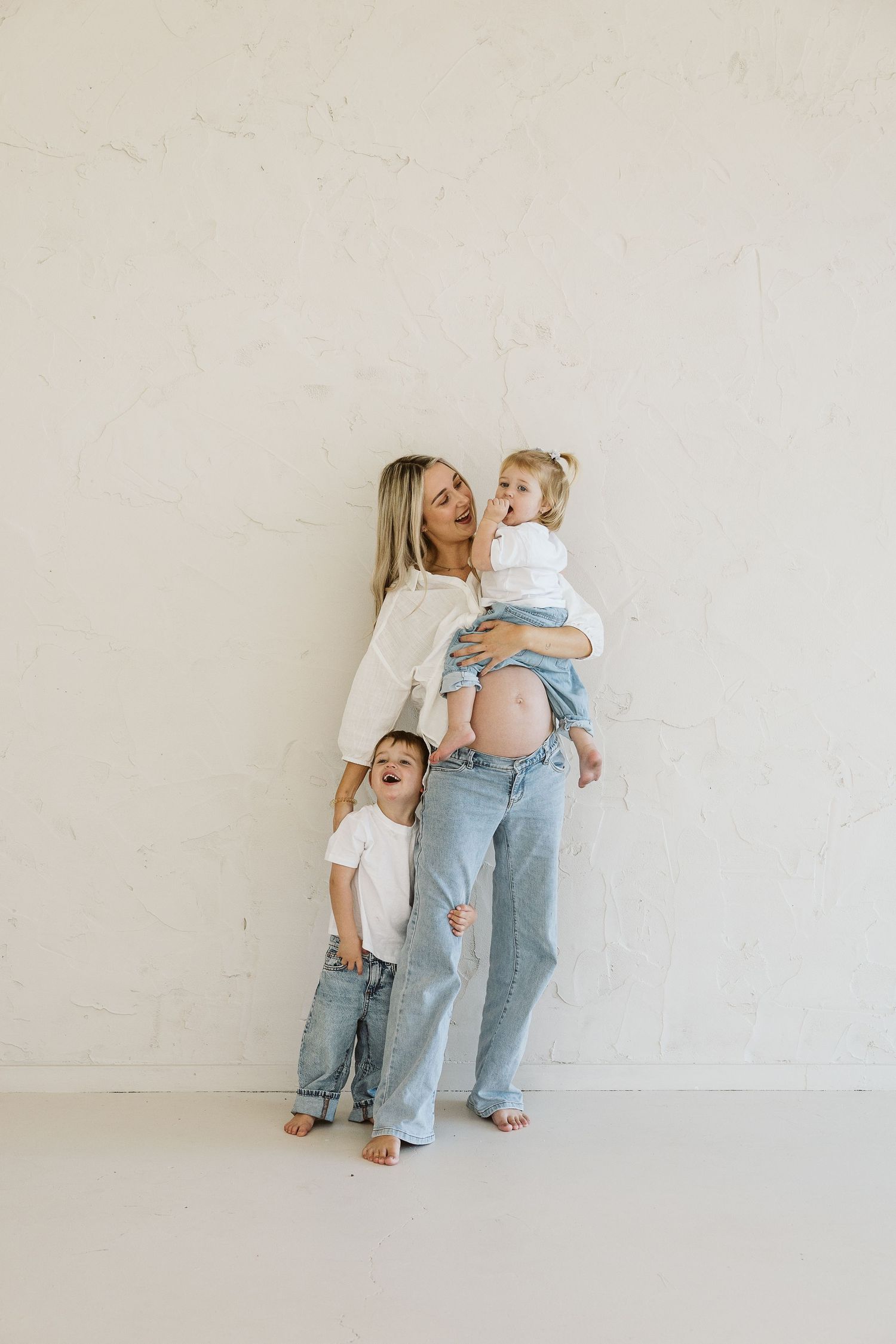 Pregnant woman in jeans holds toddler while young boy leans against her, maternity photo against white wall.