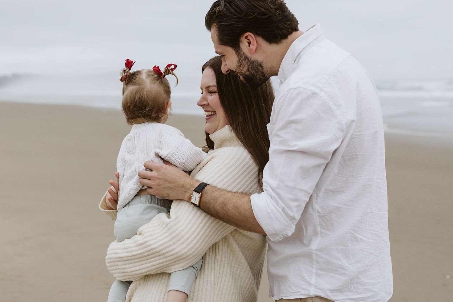 Happy young family on a beach, mother holding toddler with red bows as father leans in smiling warmly.