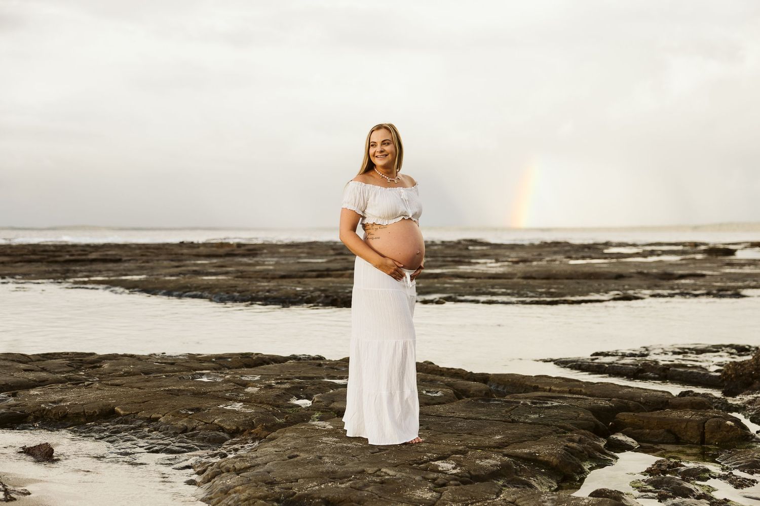 Smiling pregnant woman in white off-shoulder dress stands on rocky beach holding baby bump with rainbow sky.