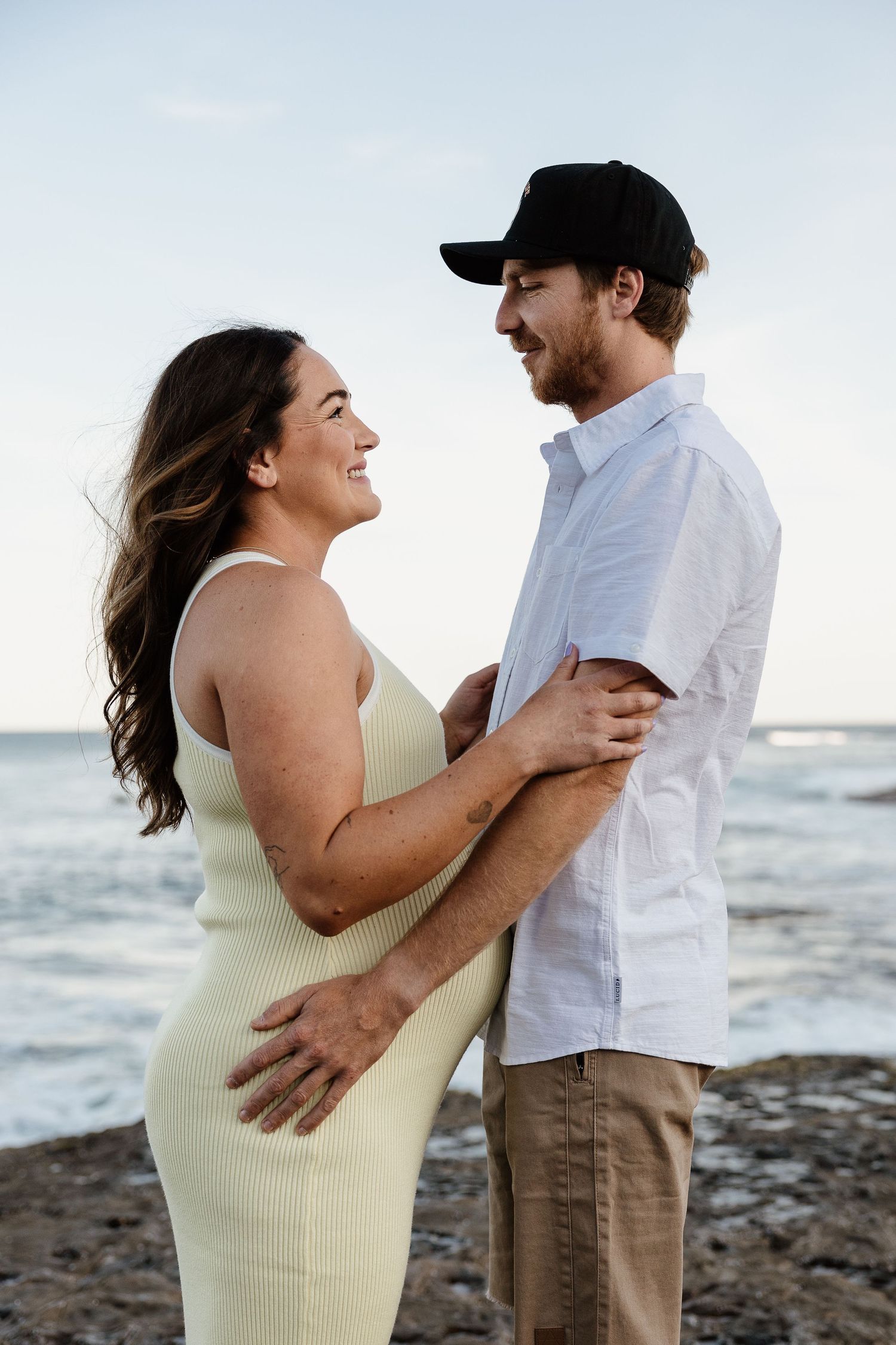 Happy couple embraces and smiles at each other on rocky ocean shoreline.