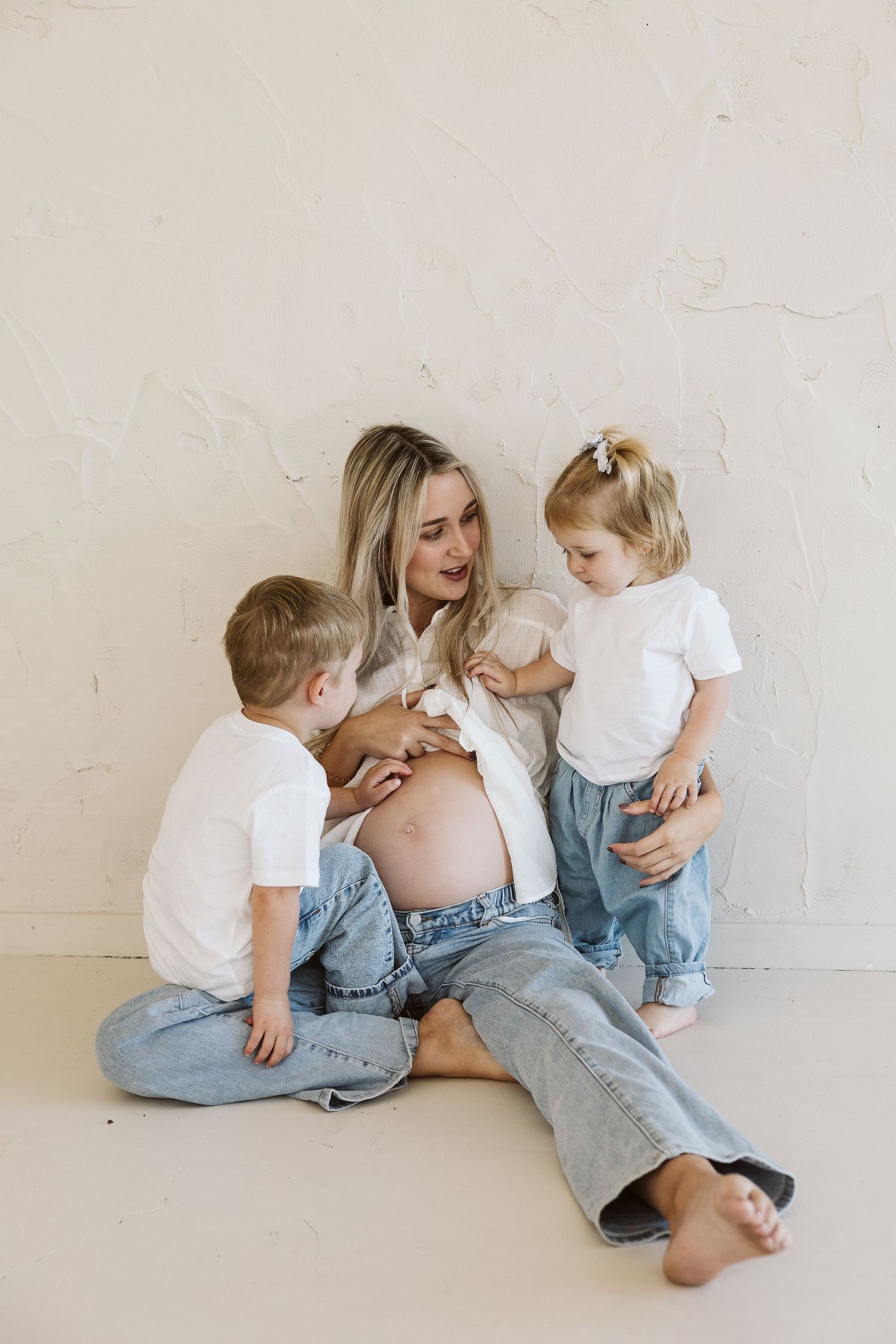 Pregnant blonde mother sitting with two young children touching her baby bump against white wall.