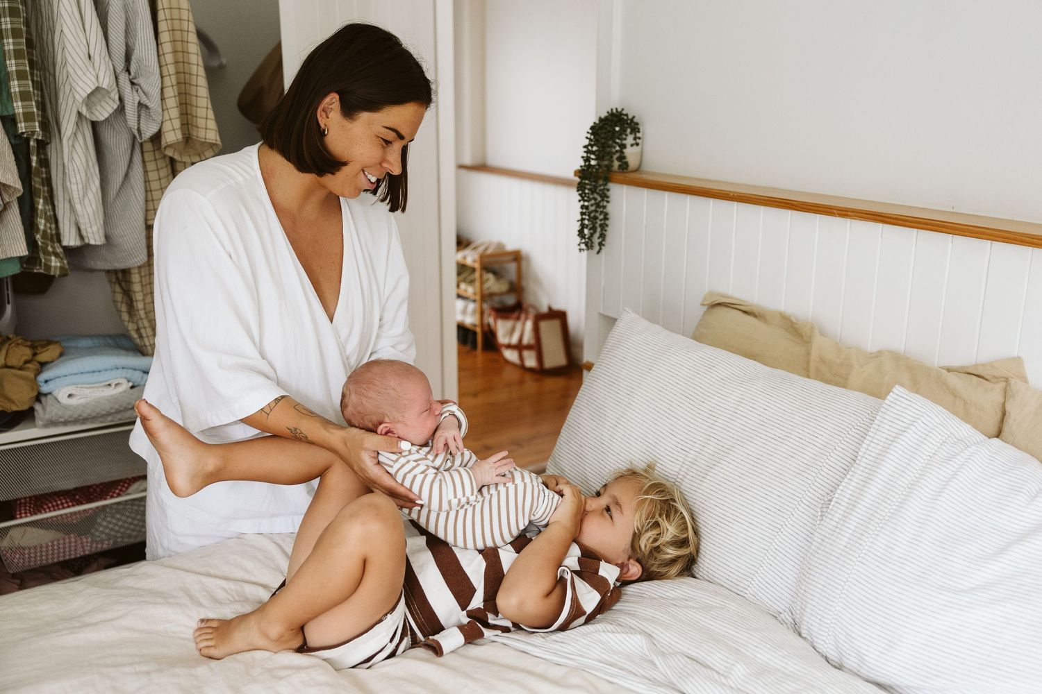 New mother in white robe sitting on bed holding newborn while young toddler lies nearby smiling.