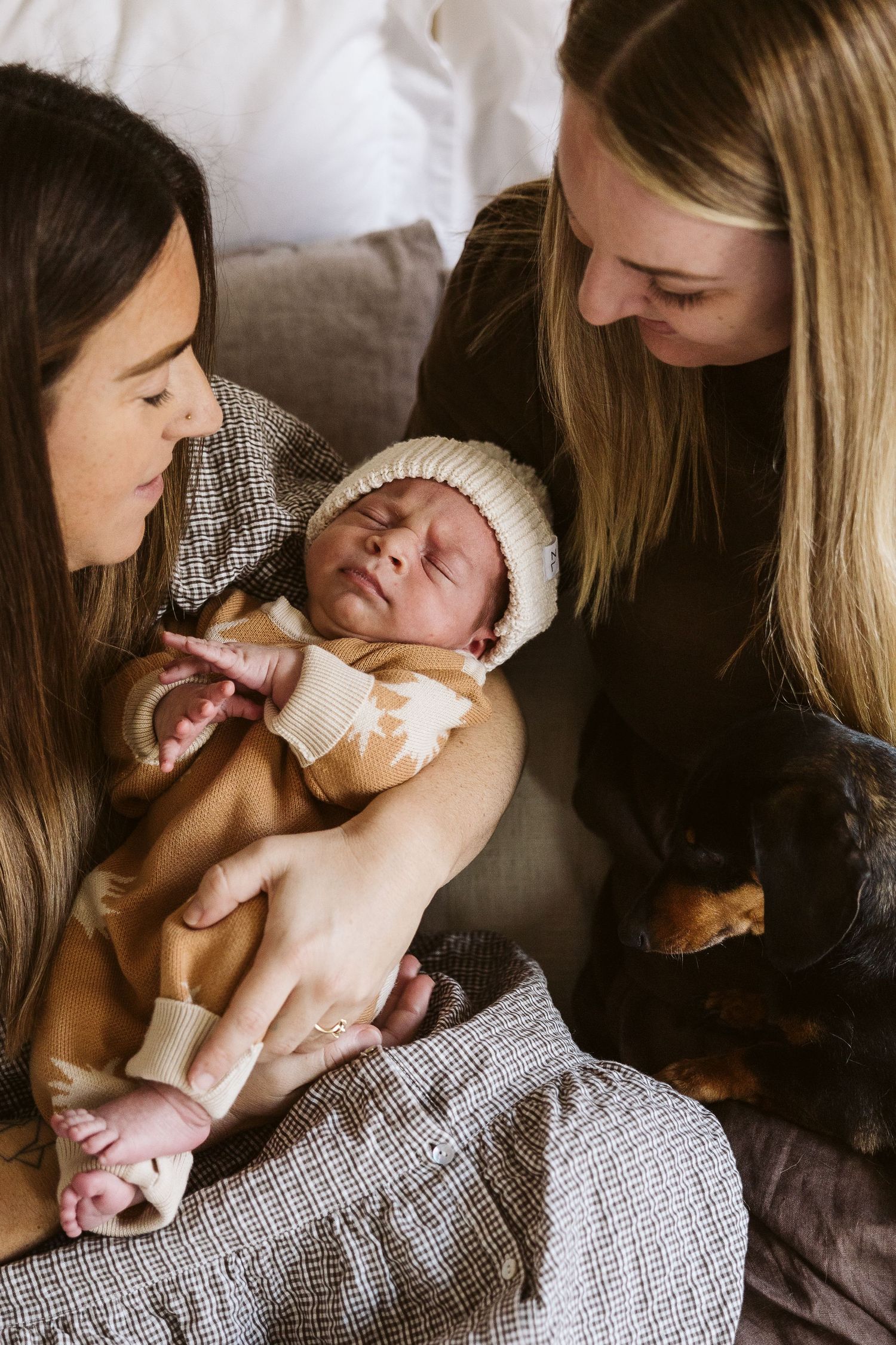 Two mothers lovingly gazing at their sleeping newborn baby dressed in tan outfit and knit hat.