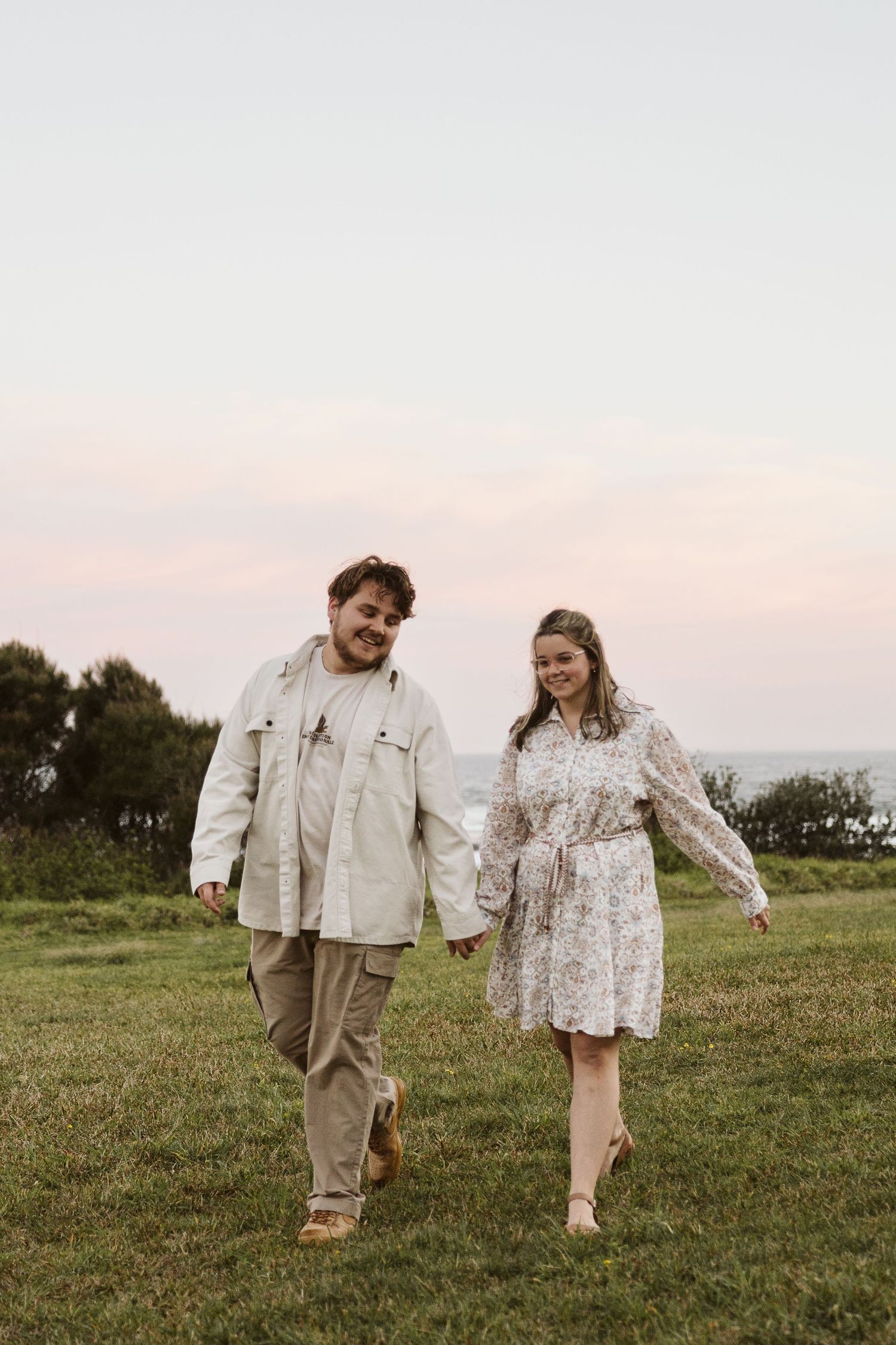 Couple walking hand in hand on green coastal lawn at dusk, both smiling and dressed in neutral tones.