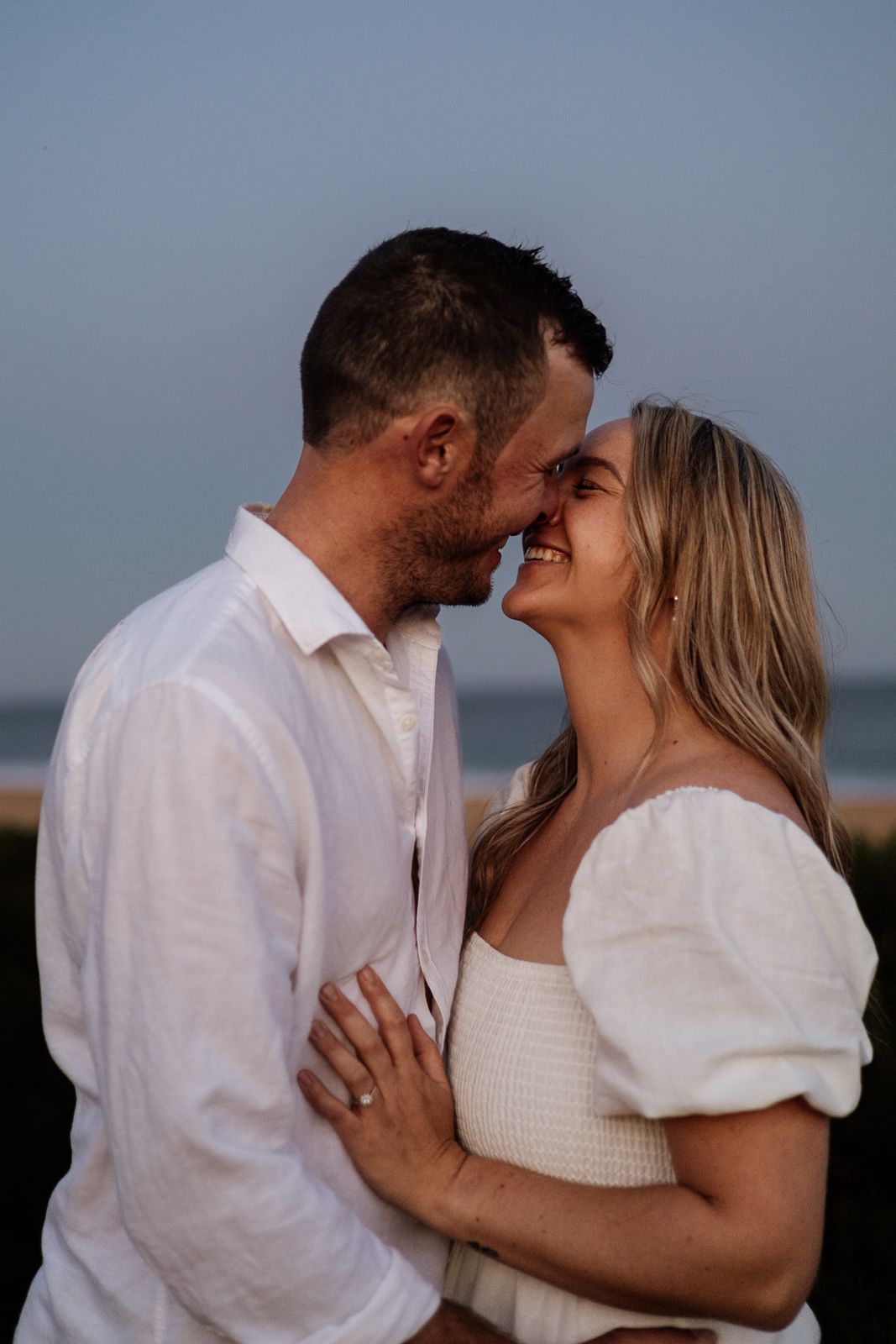 Couple about to kiss on beach at dusk, woman in white smocked dress showing engagement ring.