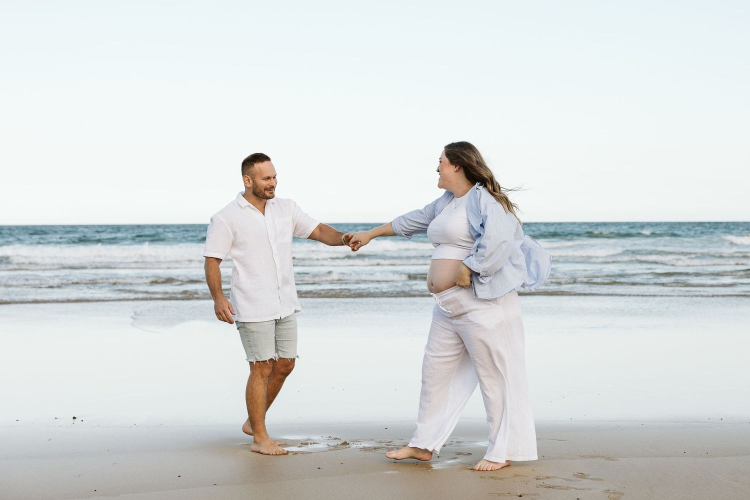 Expecting couple holding hands and dancing together on a sunny ocean beach.