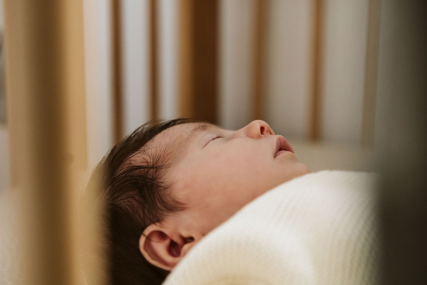 Peaceful newborn baby sleeping on their back in a wooden crib wrapped in a soft white blanket.