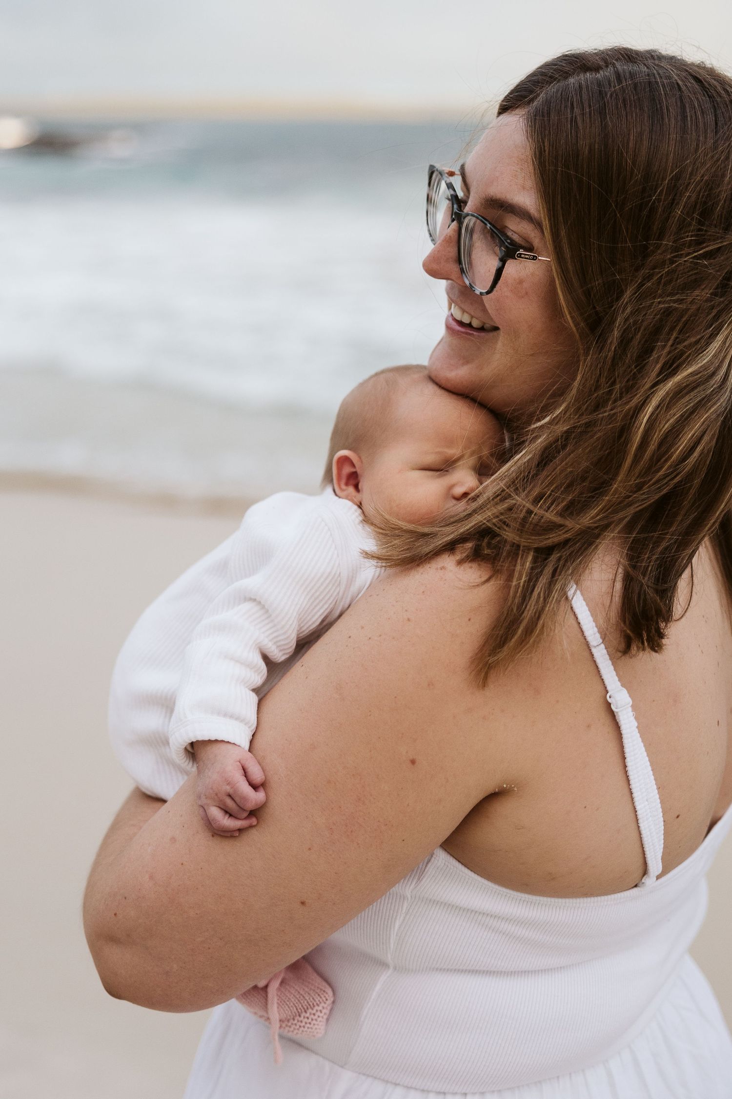Smiling mother holds sleeping newborn baby against her shoulder on sandy beach.