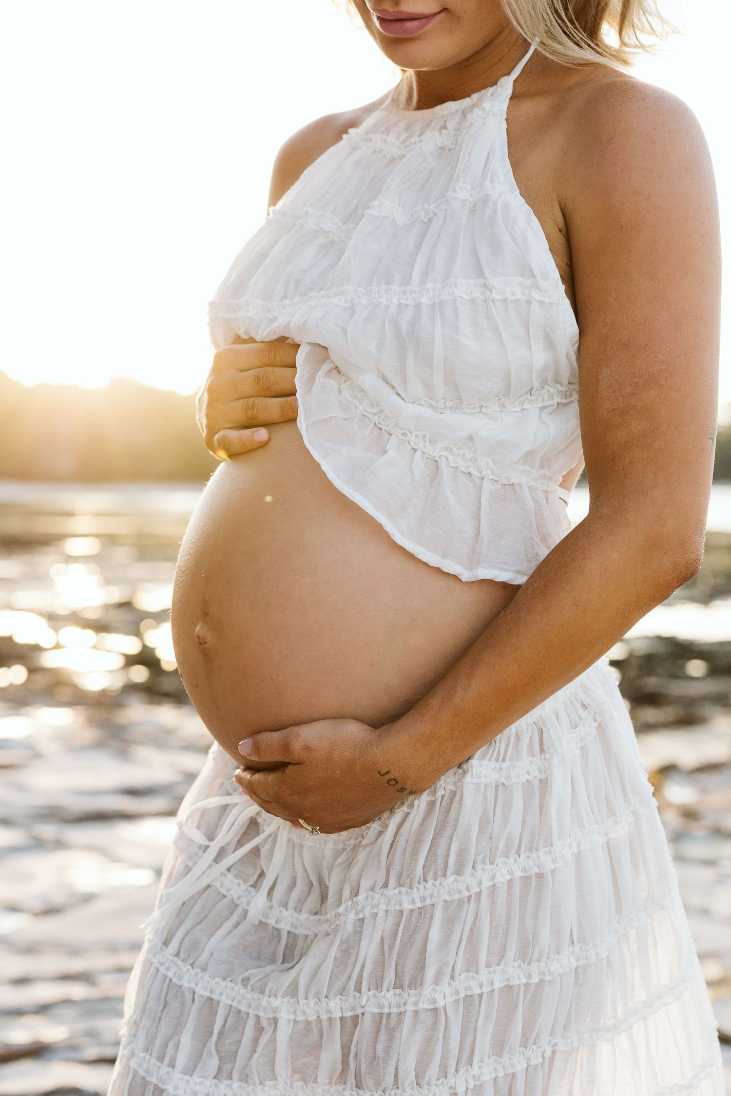 Pregnant woman in white halter dress cradling her baby bump on a sunlit beach.