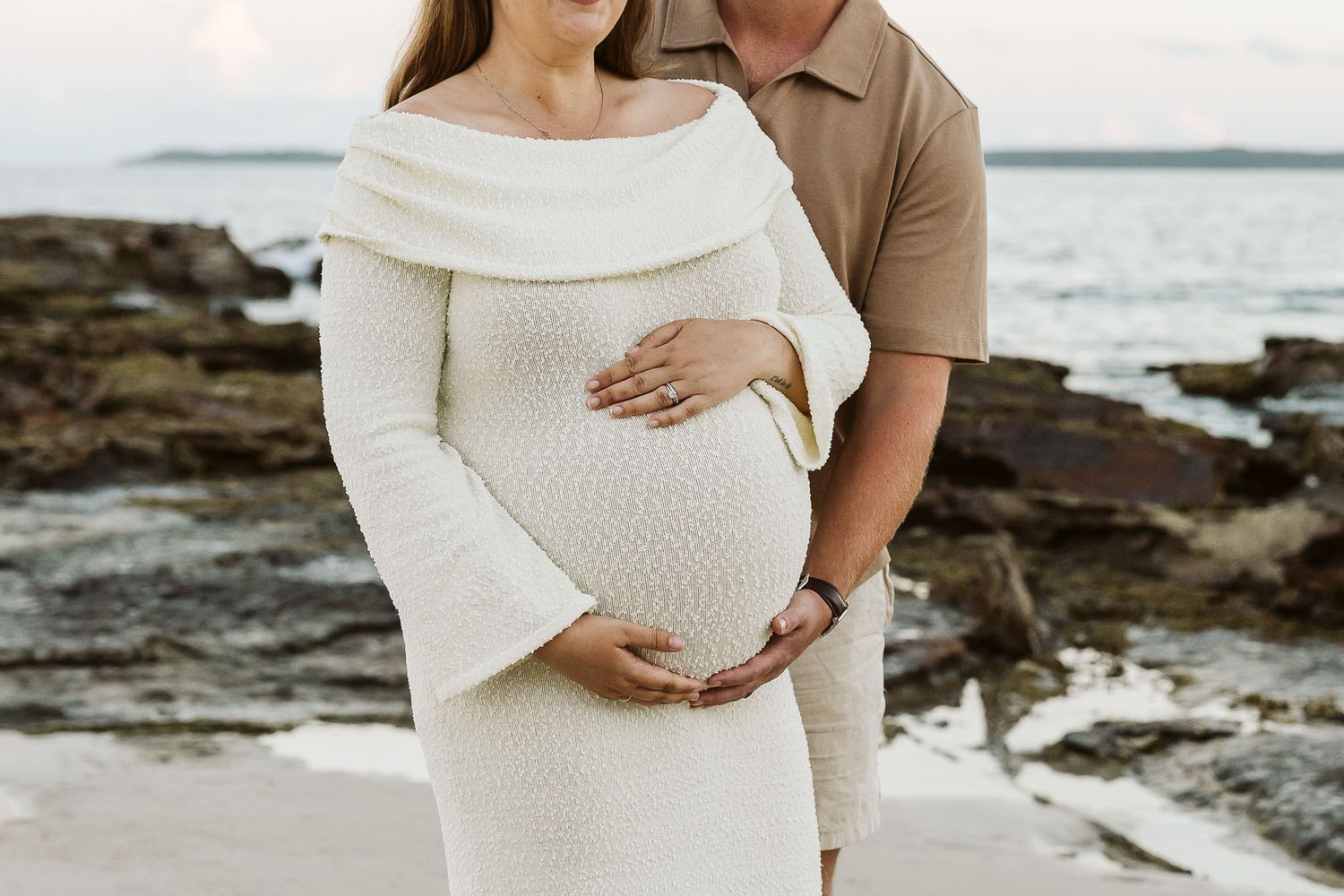 Couple on beach, man in tan shirt embraces pregnant woman in cream textured off-shoulder dress at sunset.