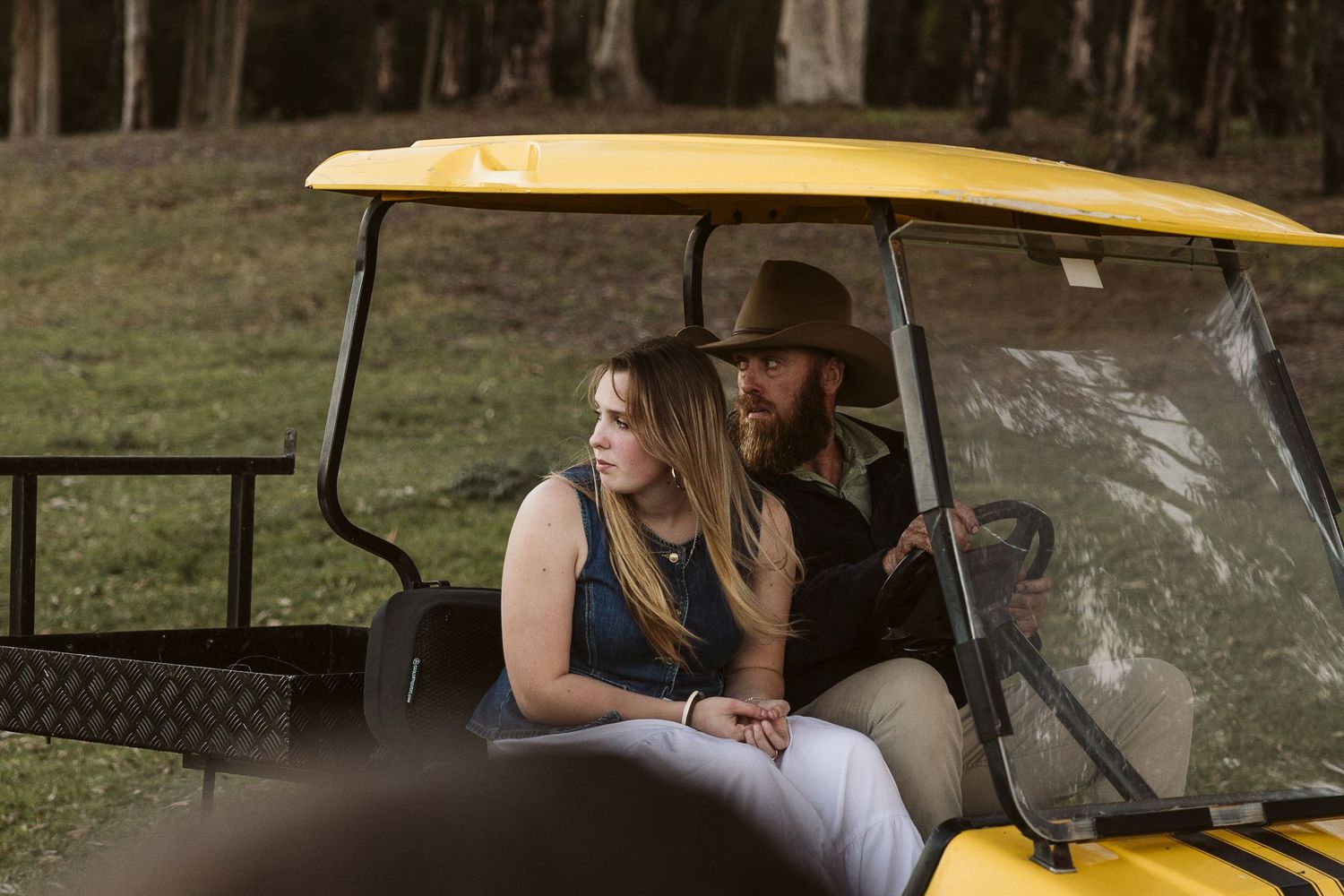 A man in a cowboy hat and woman in a blue top ride together in a yellow utility vehicle outdoors.