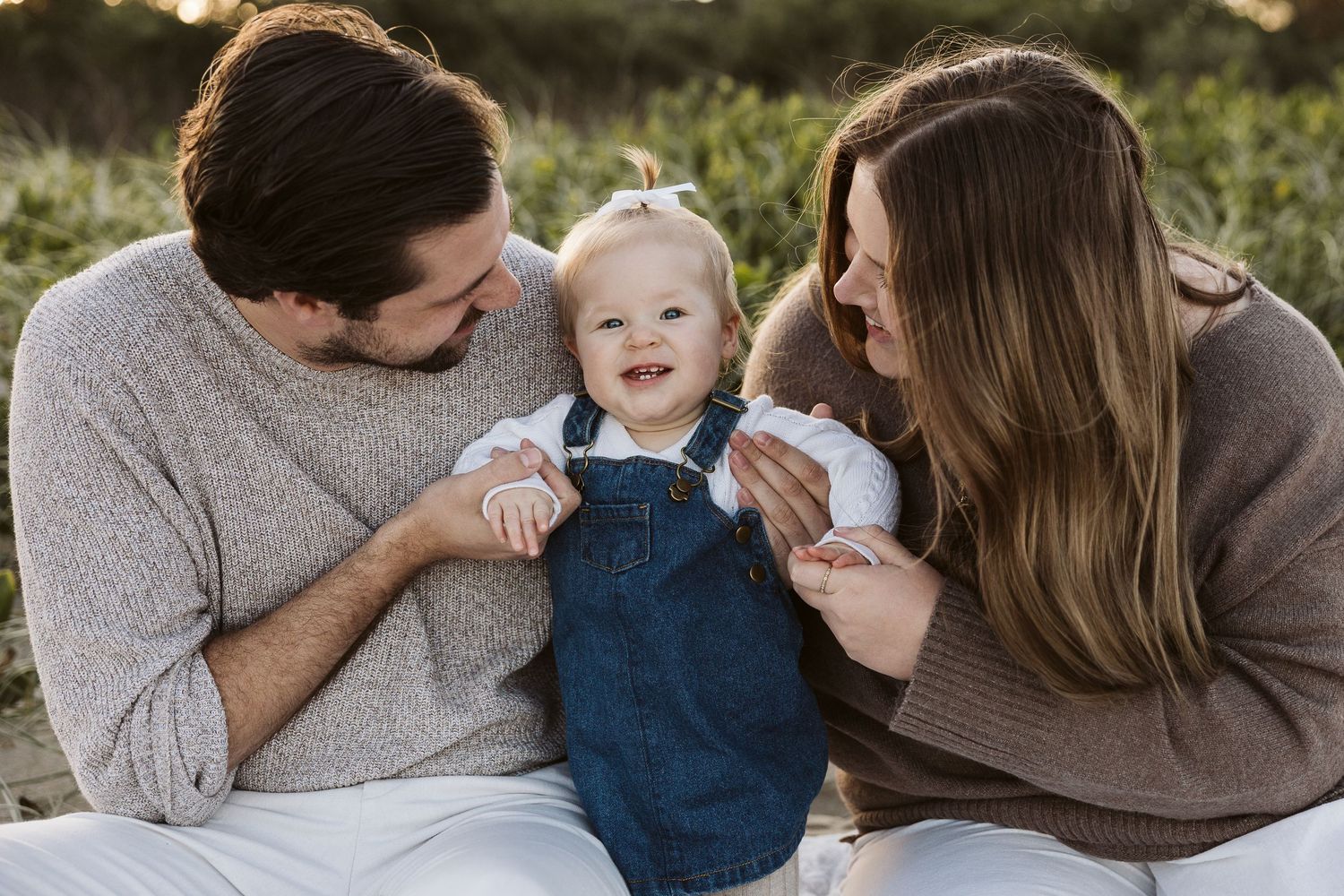 Happy baby in denim overalls smiles between loving parents during a warm outdoor family portrait session.