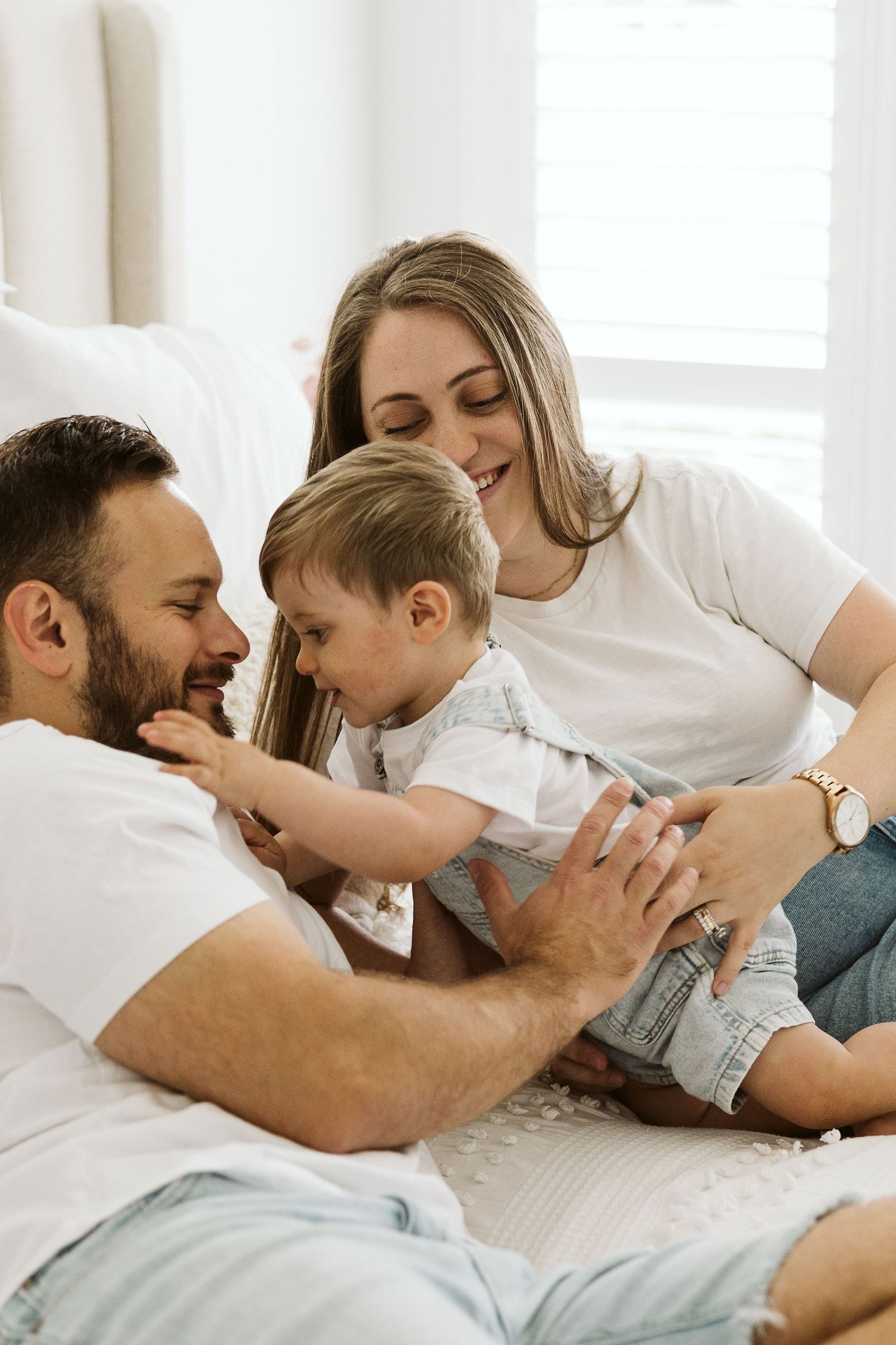 Happy family with parents and toddler son playing together on bed in bright white bedroom.