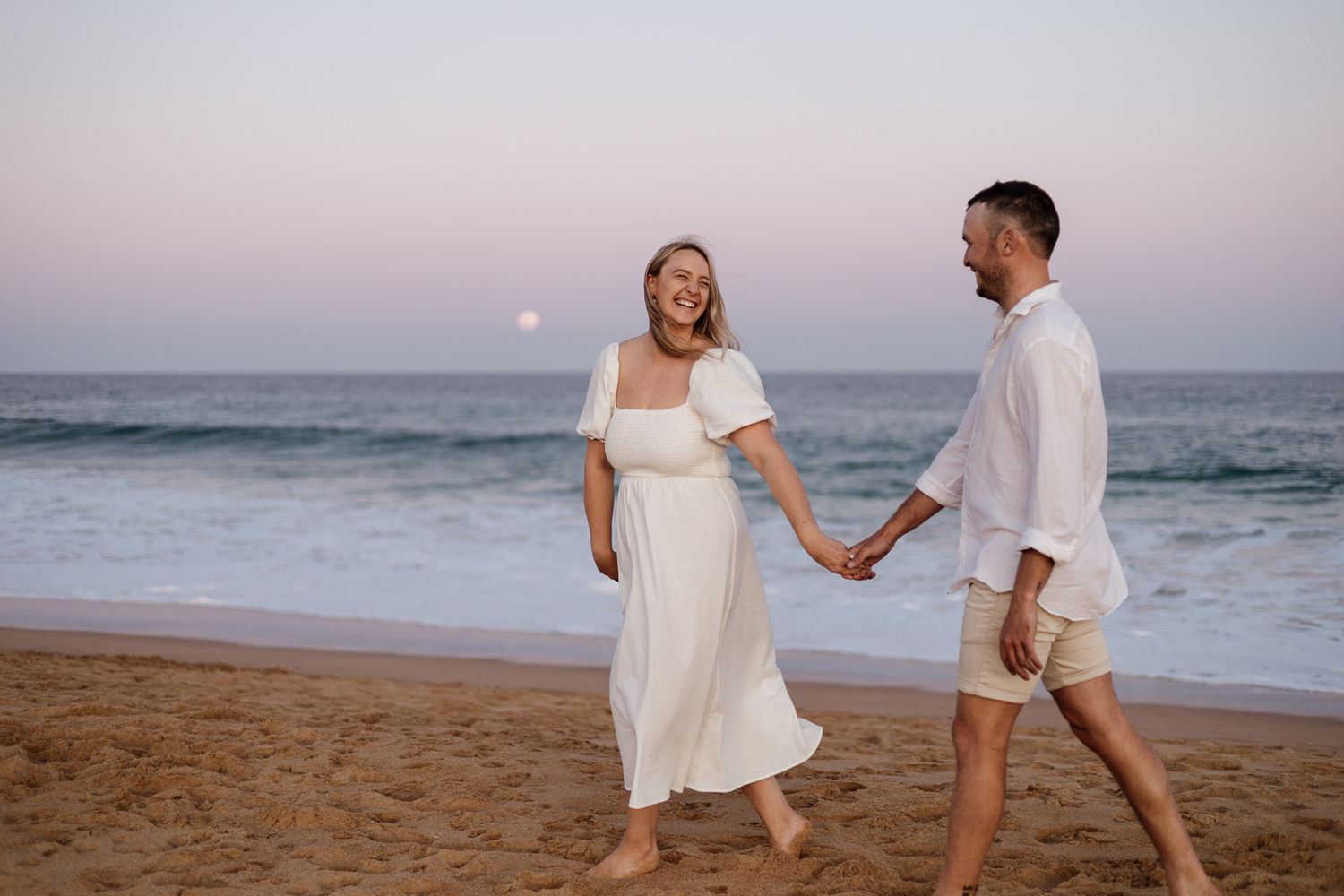 Smiling couple holding hands on sandy beach at sunset with full moon rising over ocean.