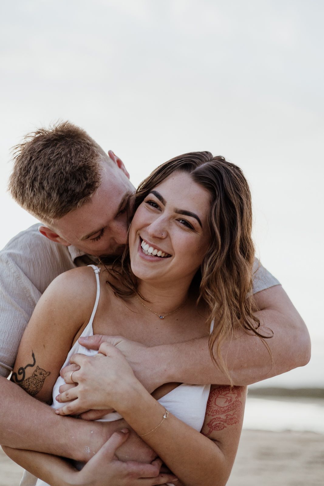 Man kissing woman on the cheek from behind as she laughs during a sunny beach engagement session.